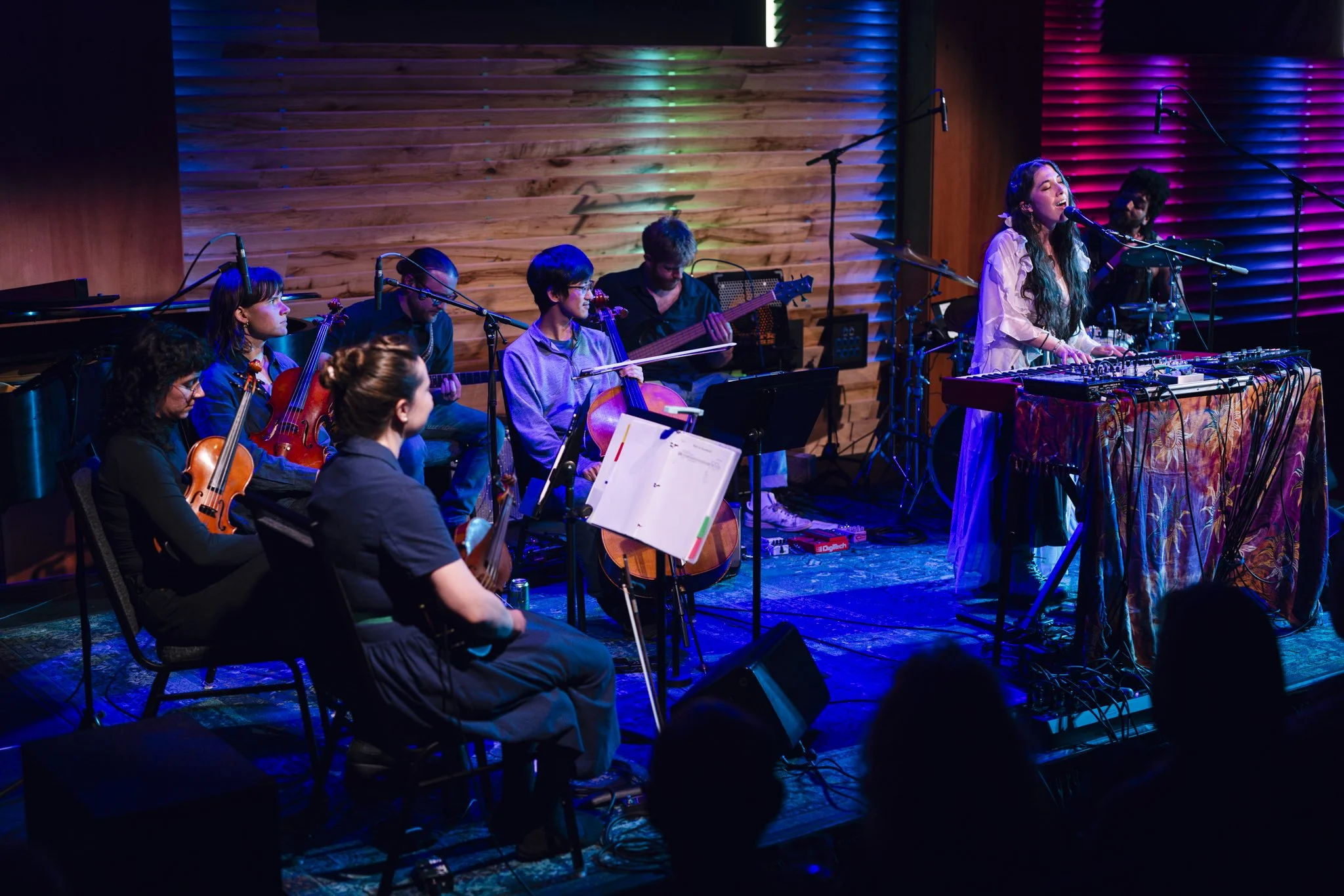 A live musical performance with a female singer at a keyboard and a band of musicians playing string and wind instruments on a stage with colorful lighting.