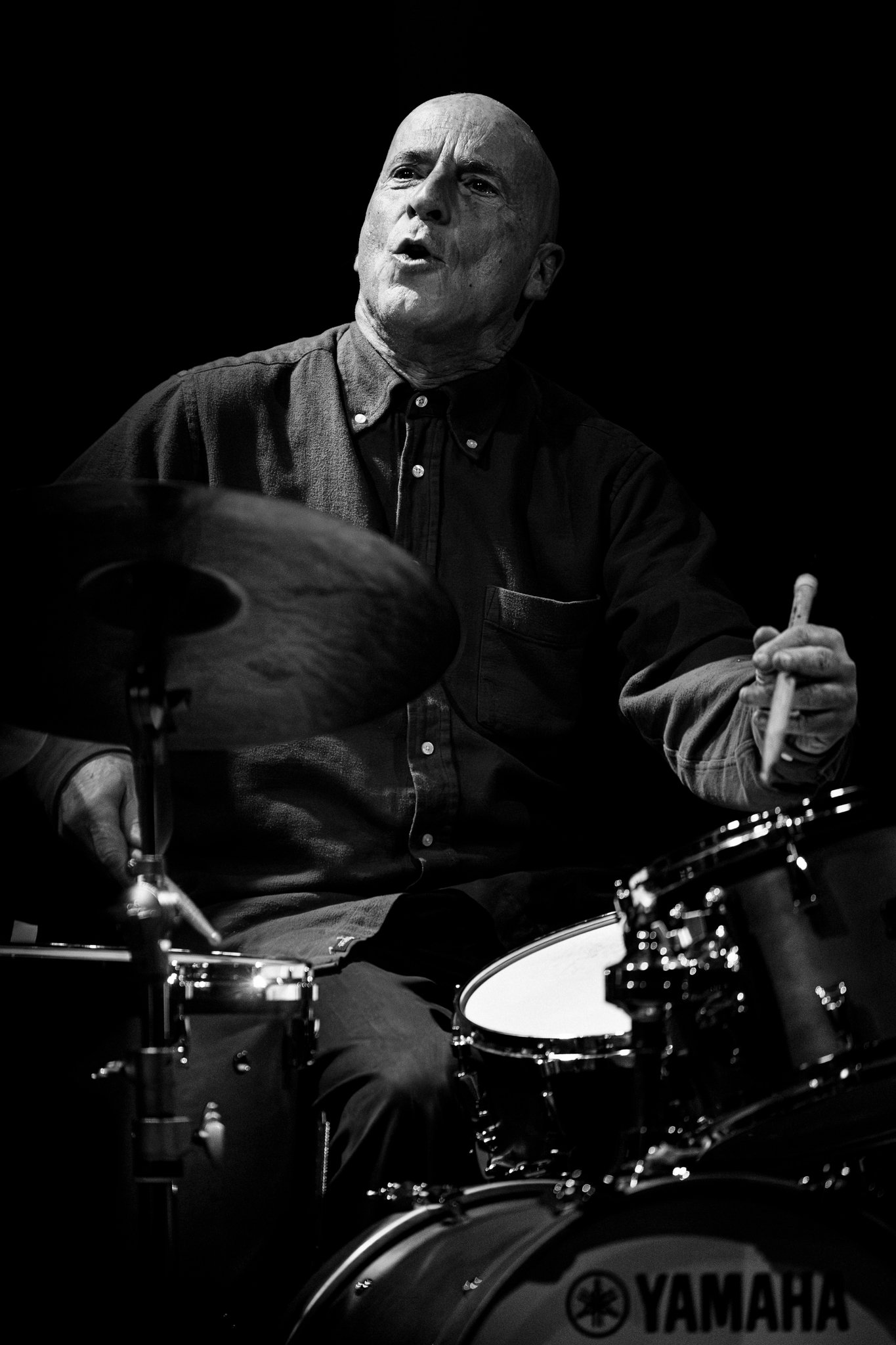 Black and white photo of a man playing the drums, holding a drumstick in his right hand, with Yamaha drum set visible.