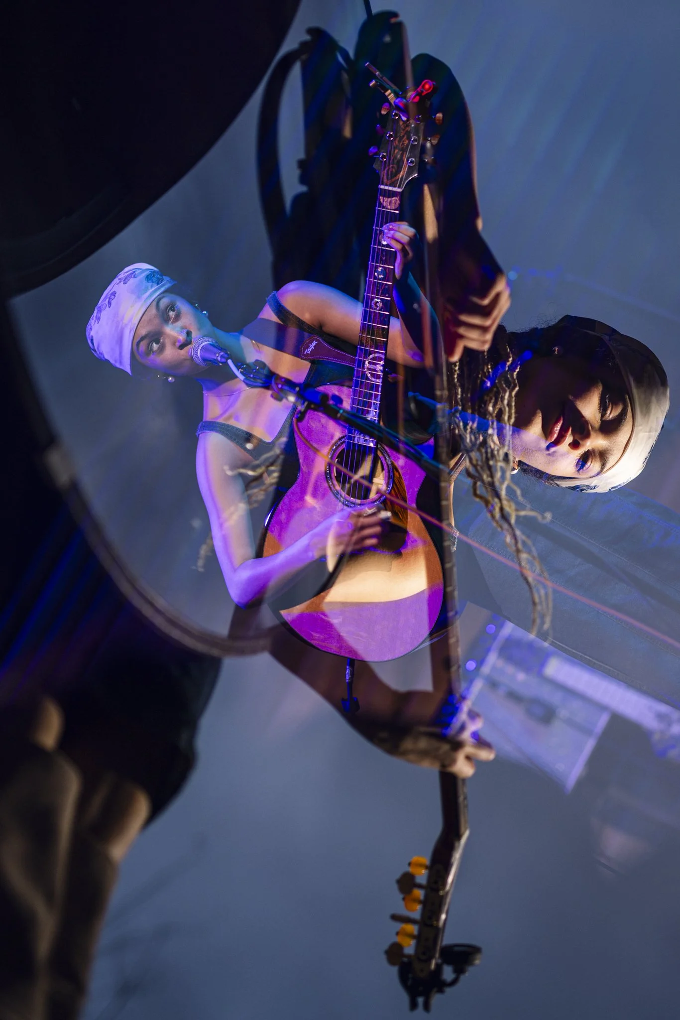 A woman with a headscarf playing an acoustic guitar and singing into a microphone, reflected in a large round mirror on a dimly lit stage.