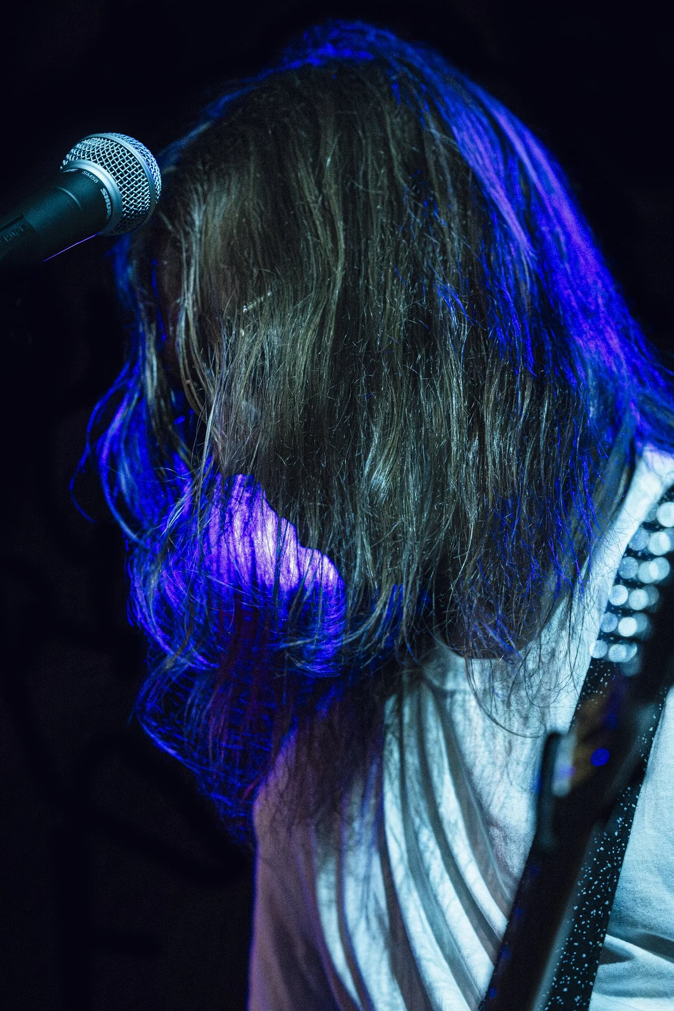 A musician with long hair down, illuminated by blue stage lights, singing into a microphone.
