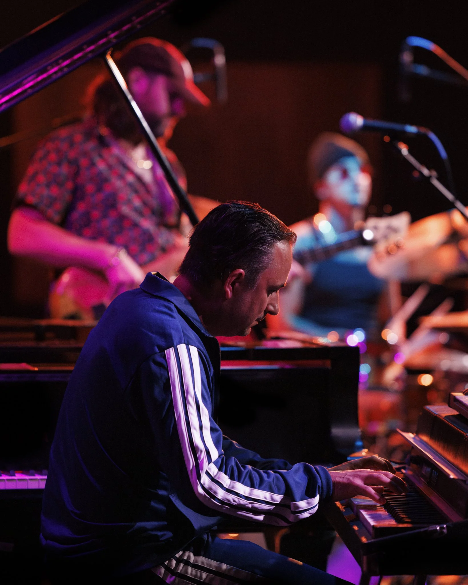 A group of musicians performing on stage, with a man playing piano in the foreground and two others playing guitar and bass in the background, illuminated by colorful stage lighting.