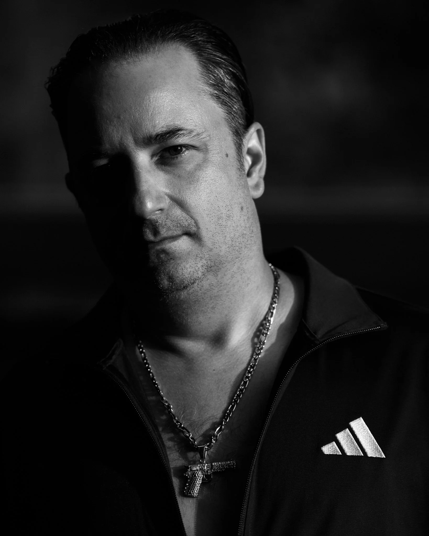 Black and white close-up portrait of a man with slicked-back hair, wearing a zippered sports jacket with a logo on the chest, and a chain necklace with a sparkly cross pendant.