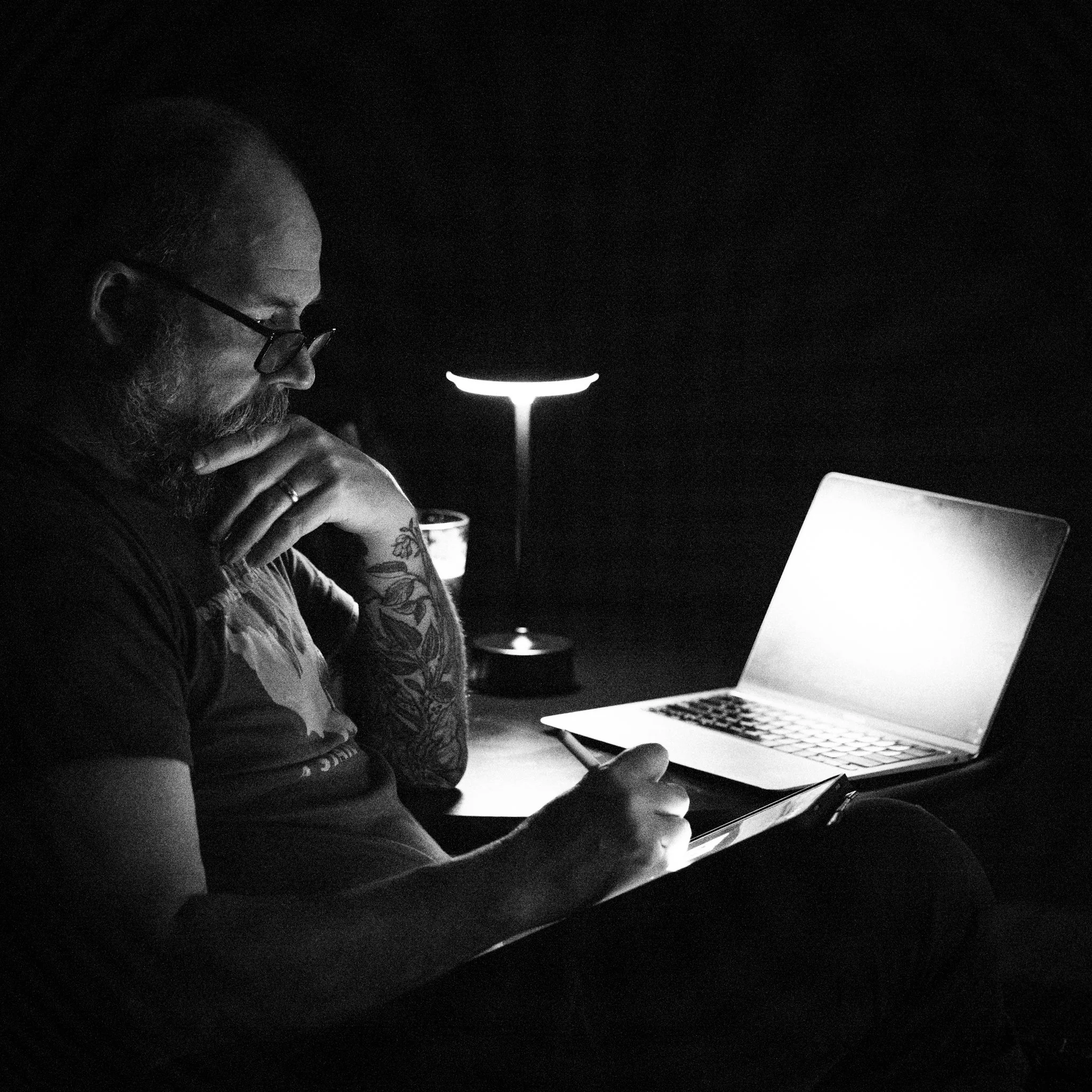 A man with glasses and a beard, sitting in a dark room, working on a tablet with a bright screen. A laptop and a small table lamp are also on the desk, providing soft light.