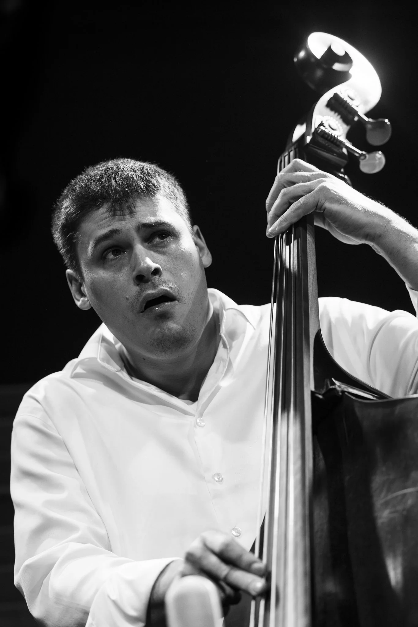Black and white photo of a young man playing a double bass, wearing a white shirt, with a focused expression.