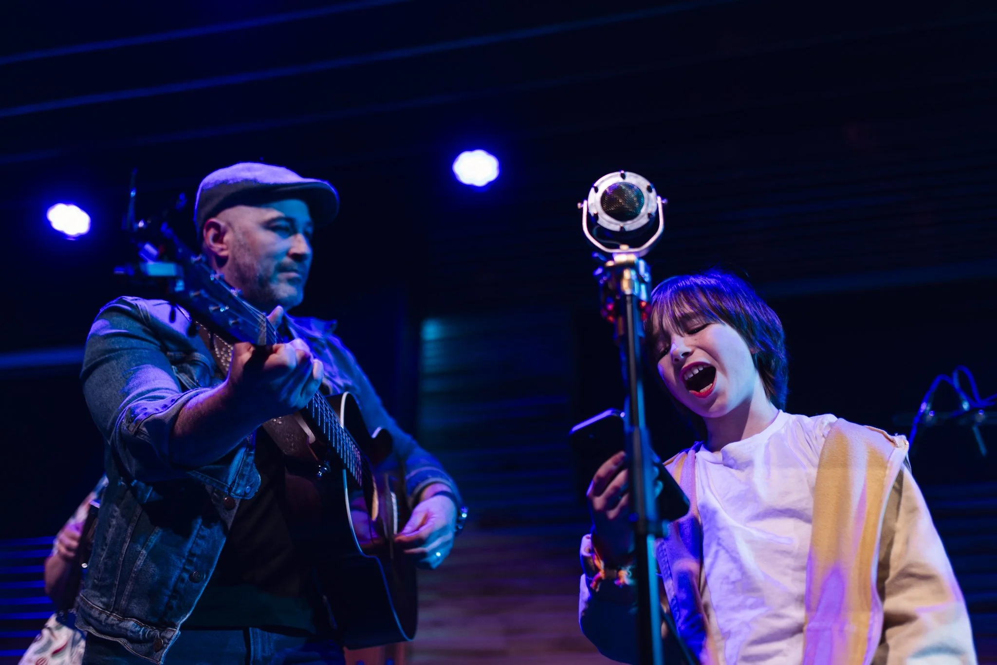 A man playing an acoustic guitar and a woman singing passionately into a microphone, both performing on stage with blue lighting.
