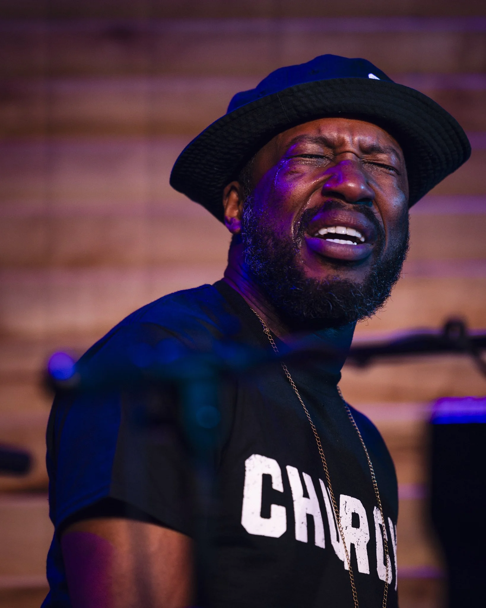 An emotional man wearing a black hat and a black shirt with white letters, crying with eyes closed, against a wooden background.