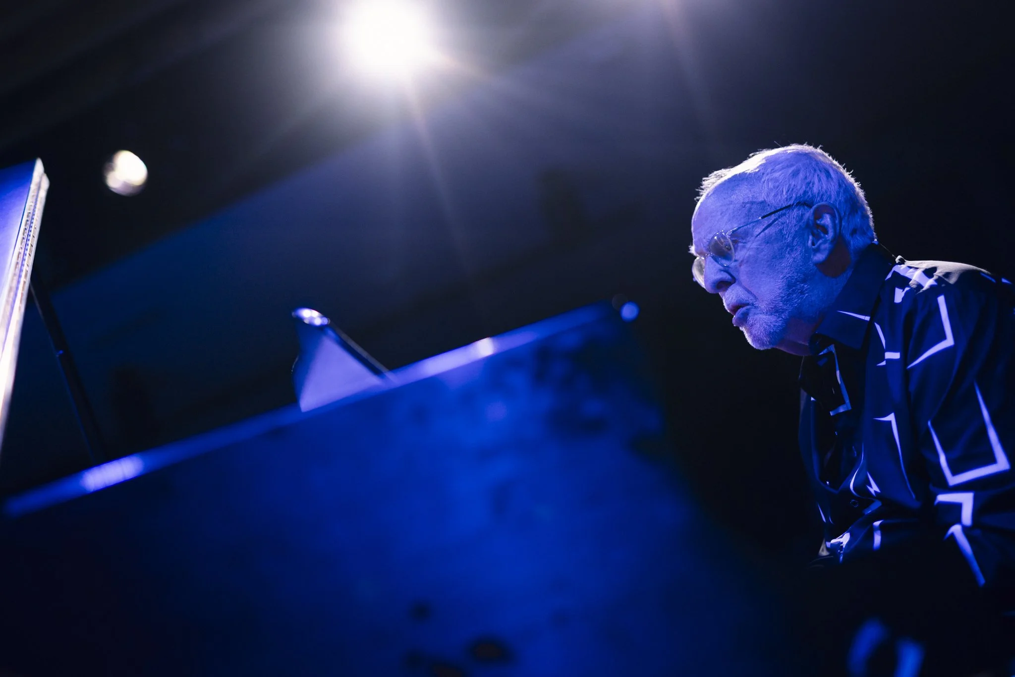 An elderly man with white hair and glasses plays the piano on stage under blue lighting, with a bright spotlight shining from above.