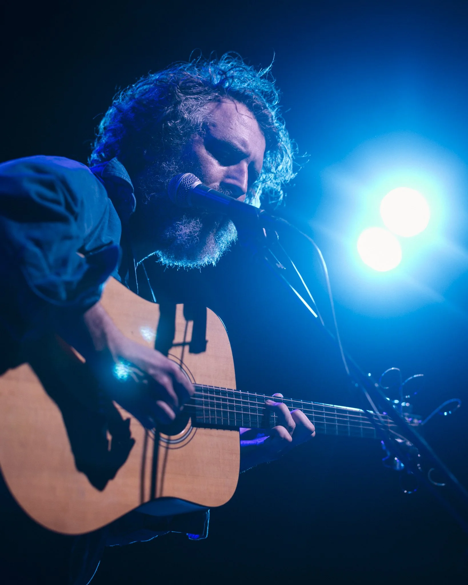 A man with curly hair and a beard playing an acoustic guitar on stage, with bright blue stage lights behind him.