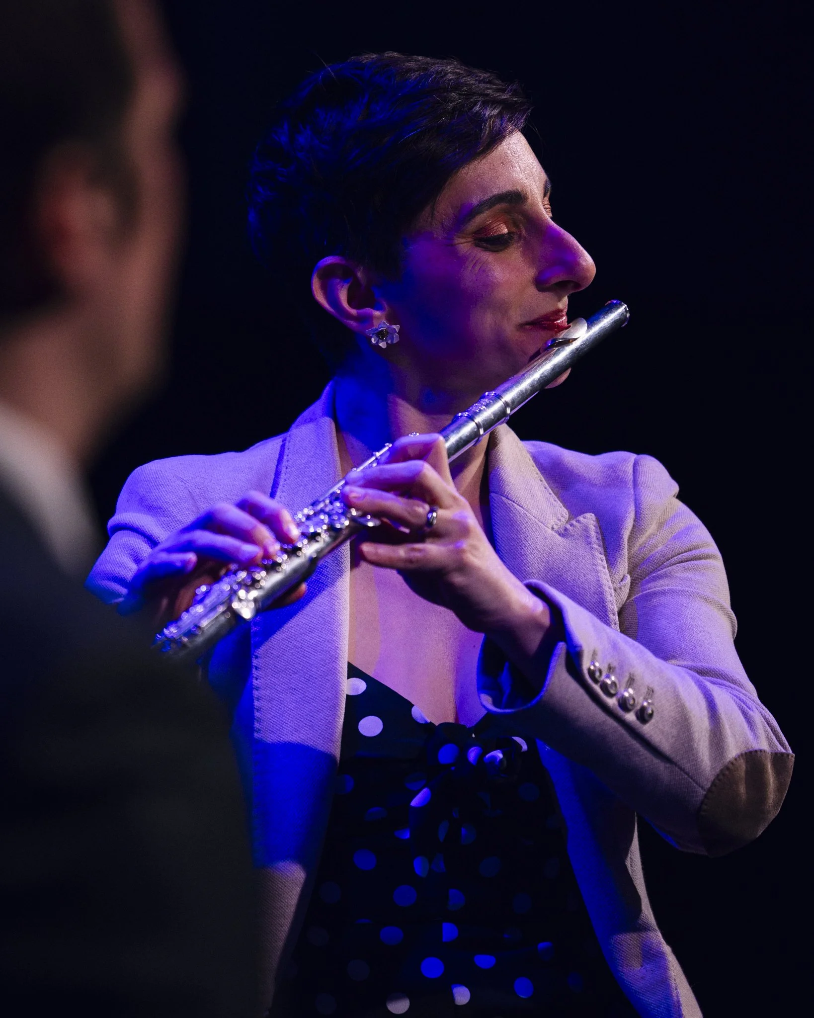 A woman with short dark hair and earrings plays a silver flute during a performance, with a blurred man in the foreground.