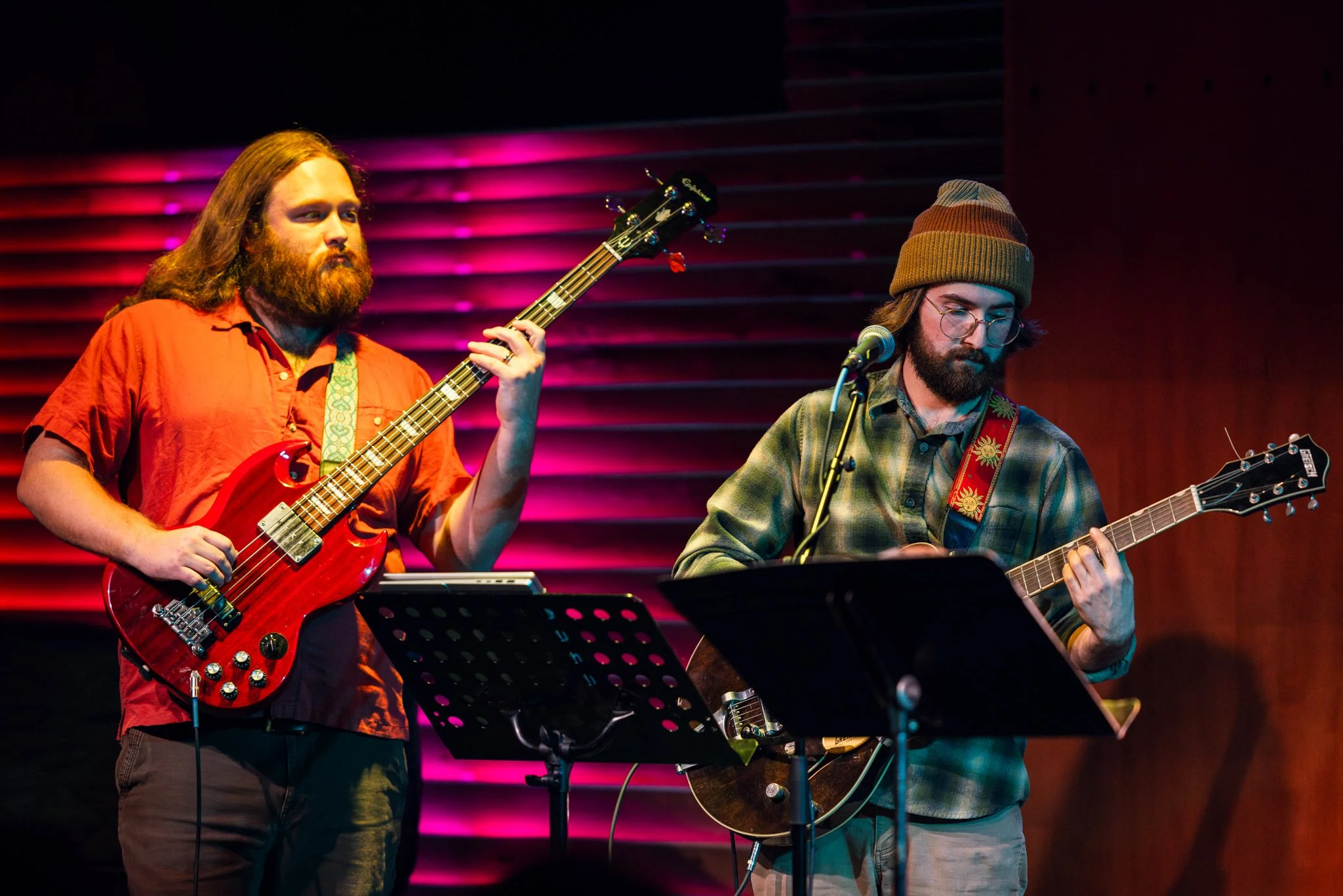 Two male musicians perform on stage, one playing a red electric bass guitar and the other playing an acoustic guitar, with music stands in front of them. The background features pink and purple lighting.
