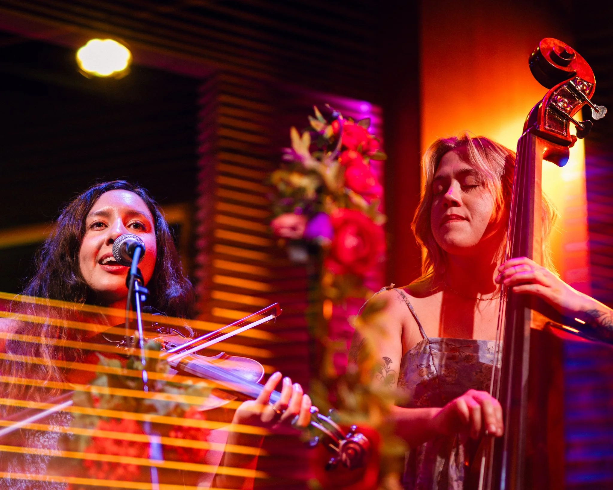 Two women performing music on stage with colorful lighting. The woman on the left is singing into a microphone, and the woman on the right is playing a cello with her eyes closed.