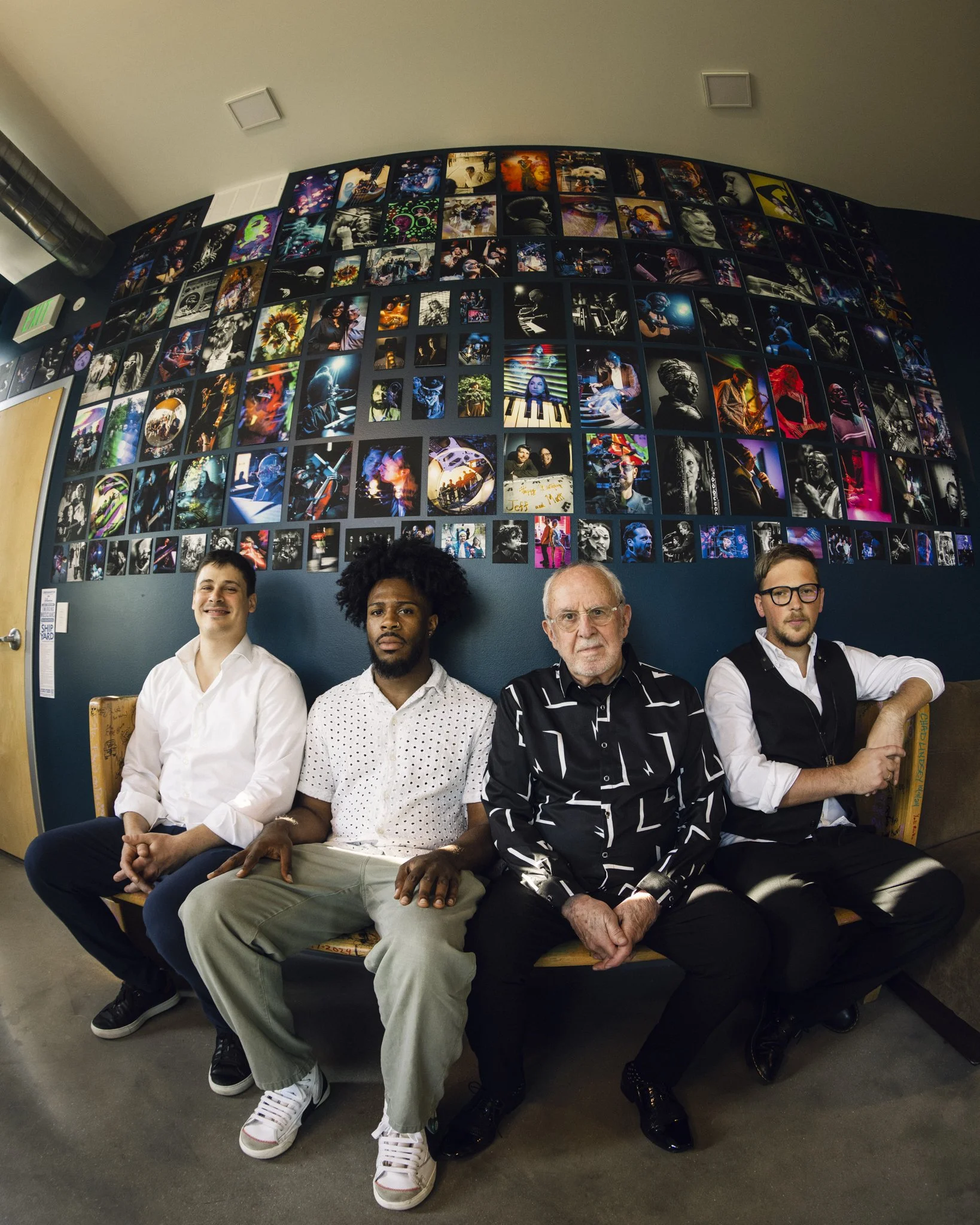 Four men sitting on a wooden bench in front of a large wall collage of colorful photographs.