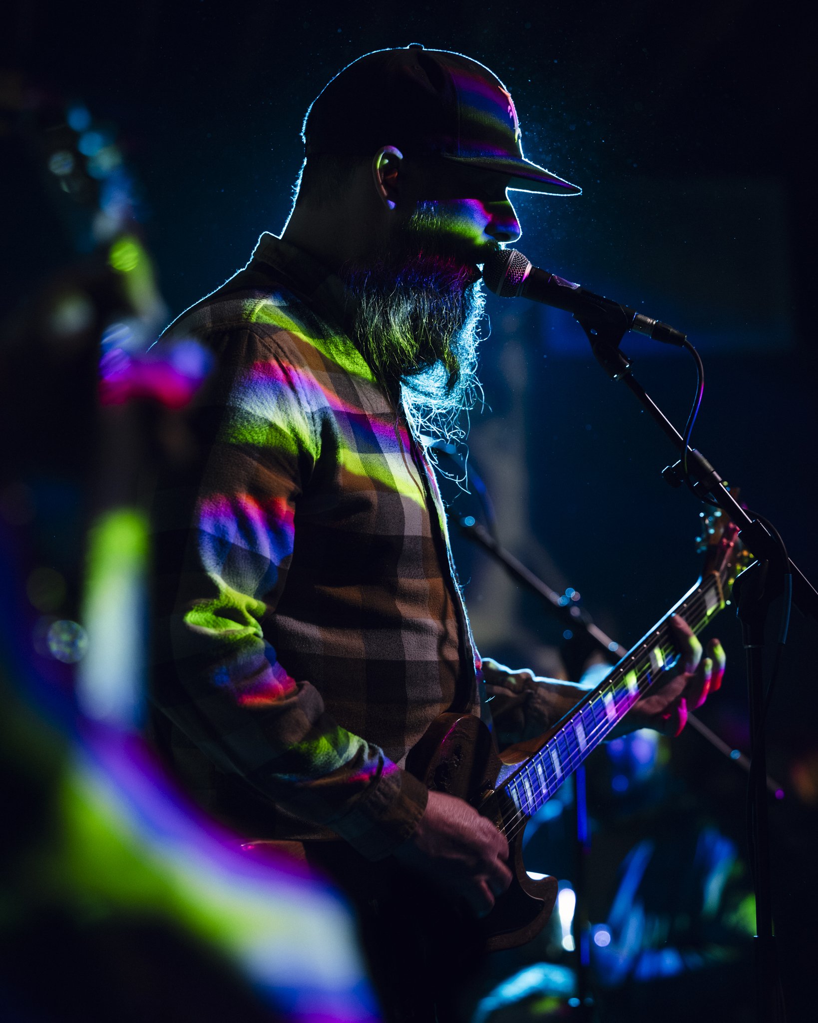 A man with a beard wearing a baseball cap and plaid shirt playing an electric guitar and singing into a microphone on stage, illuminated by colorful stage lights.