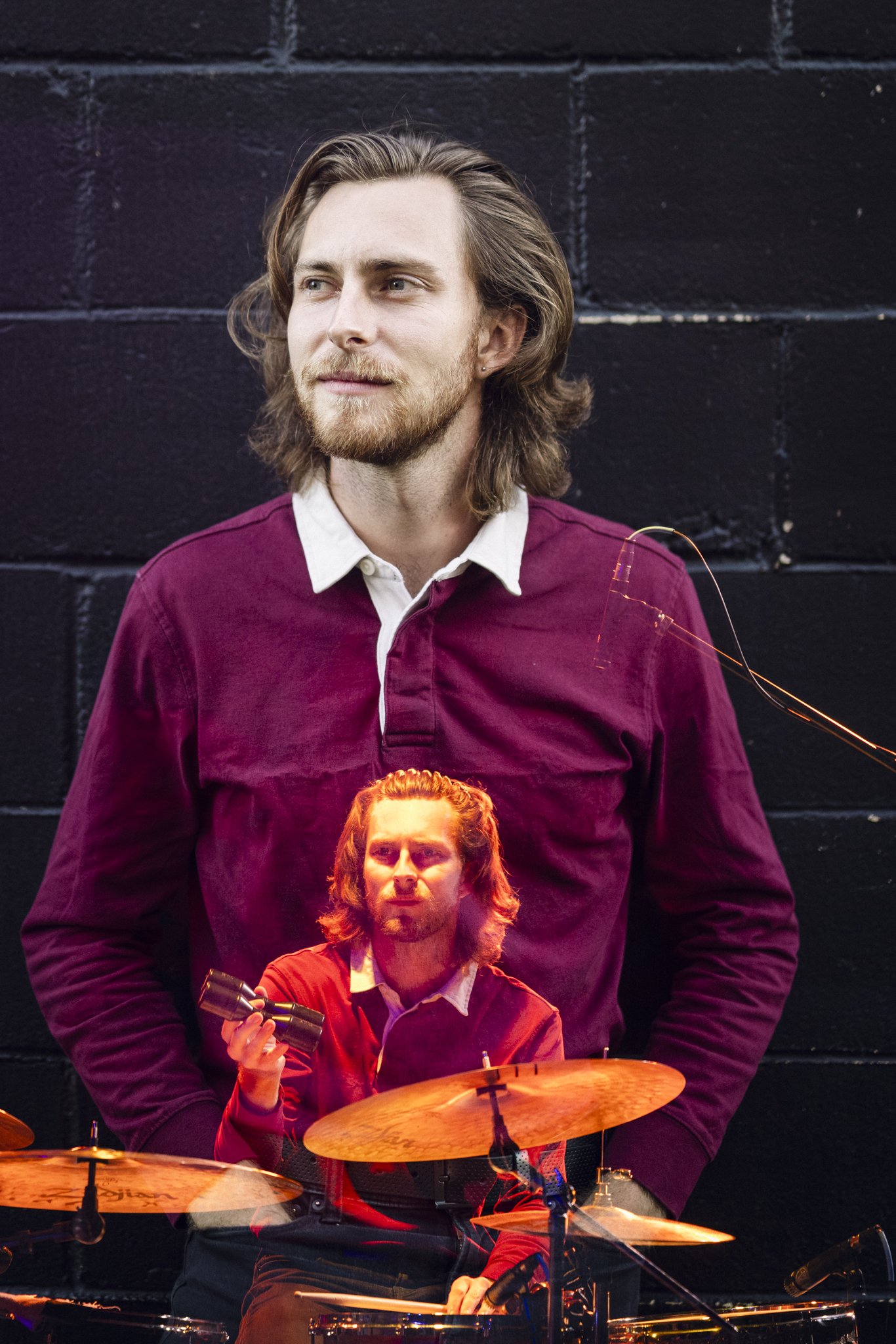 A man with long, wavy hair and a beard, wearing a maroon shirt, standing in front of a black wall.