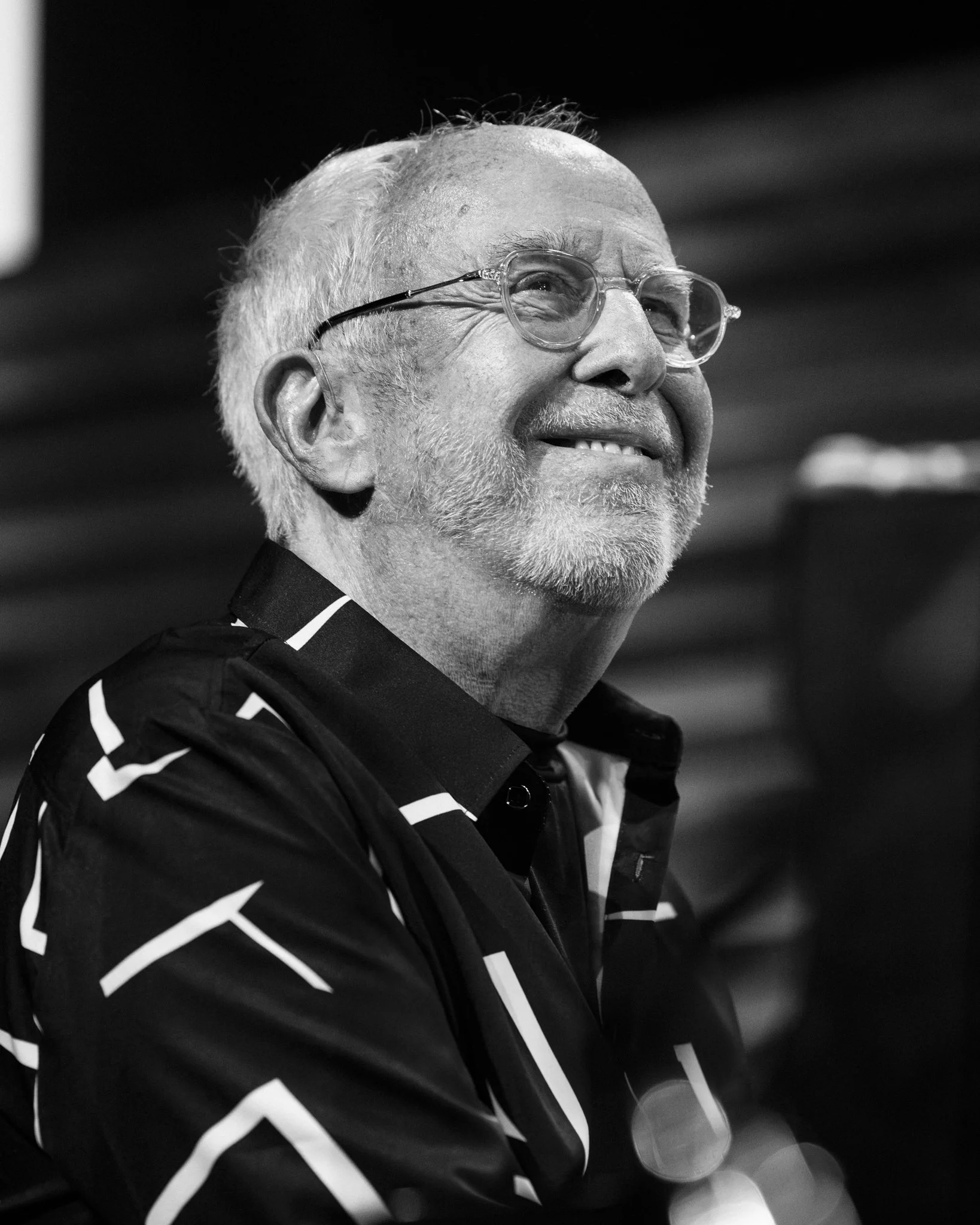 A smiling older man with glasses, white hair, and a beard, wearing a dark patterned shirt.
