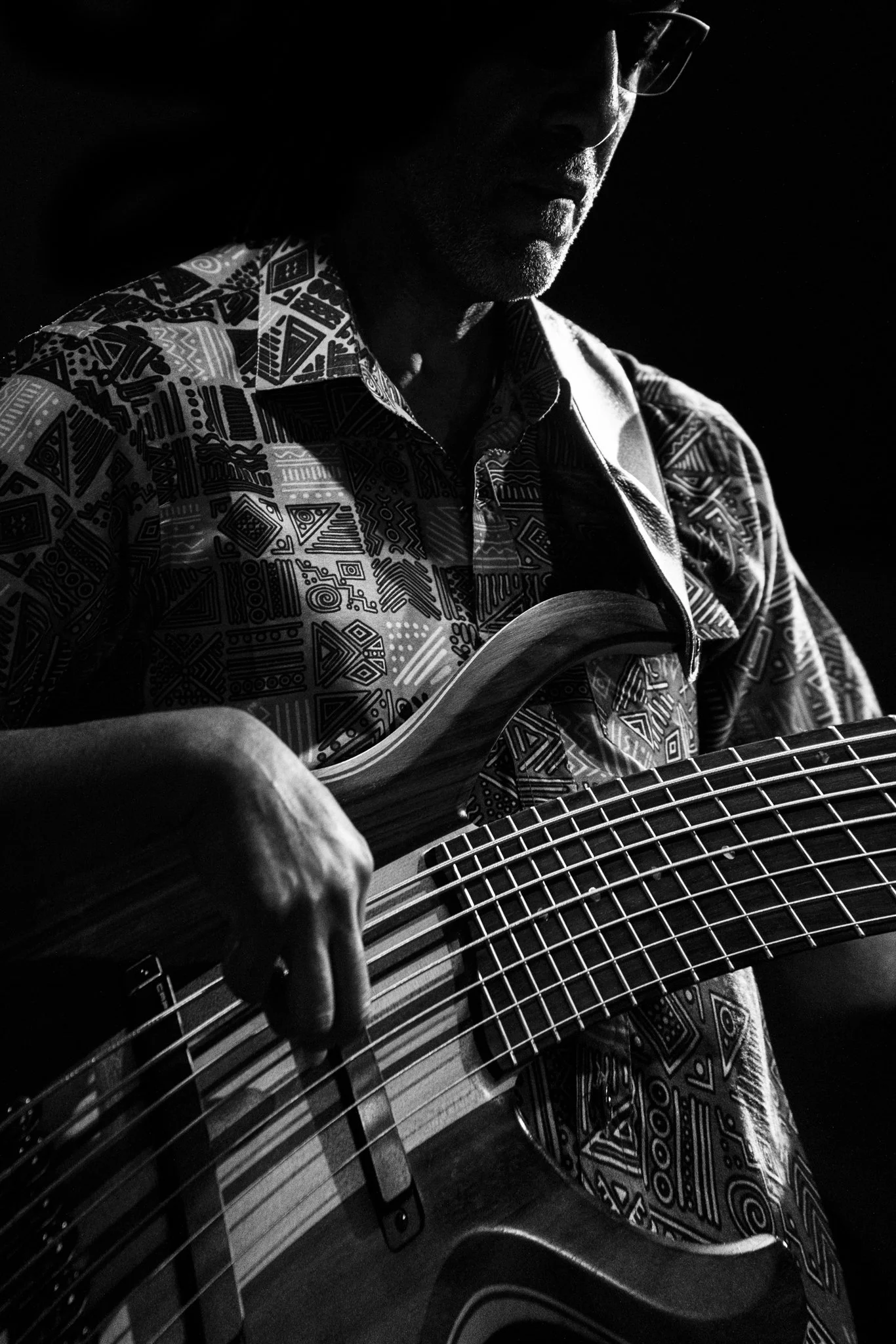 Man playing an electric bass guitar in a dark setting, wearing glasses and a patterned shirt.