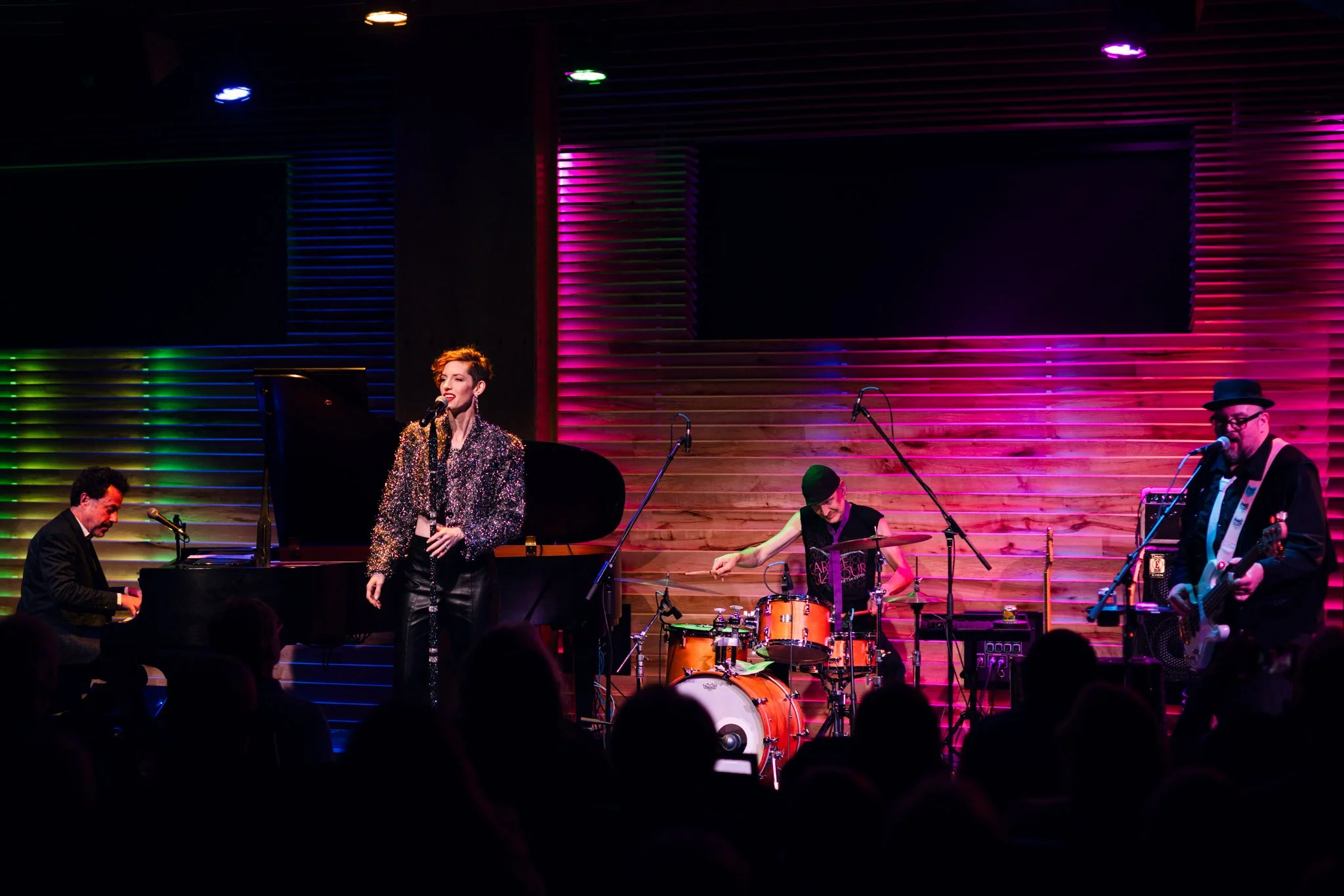 A four-piece band performs on stage with colorful lighting, with a woman singing at the center, a pianist on the left, a drummer behind her, and a guitarist on the right, in front of an audience.