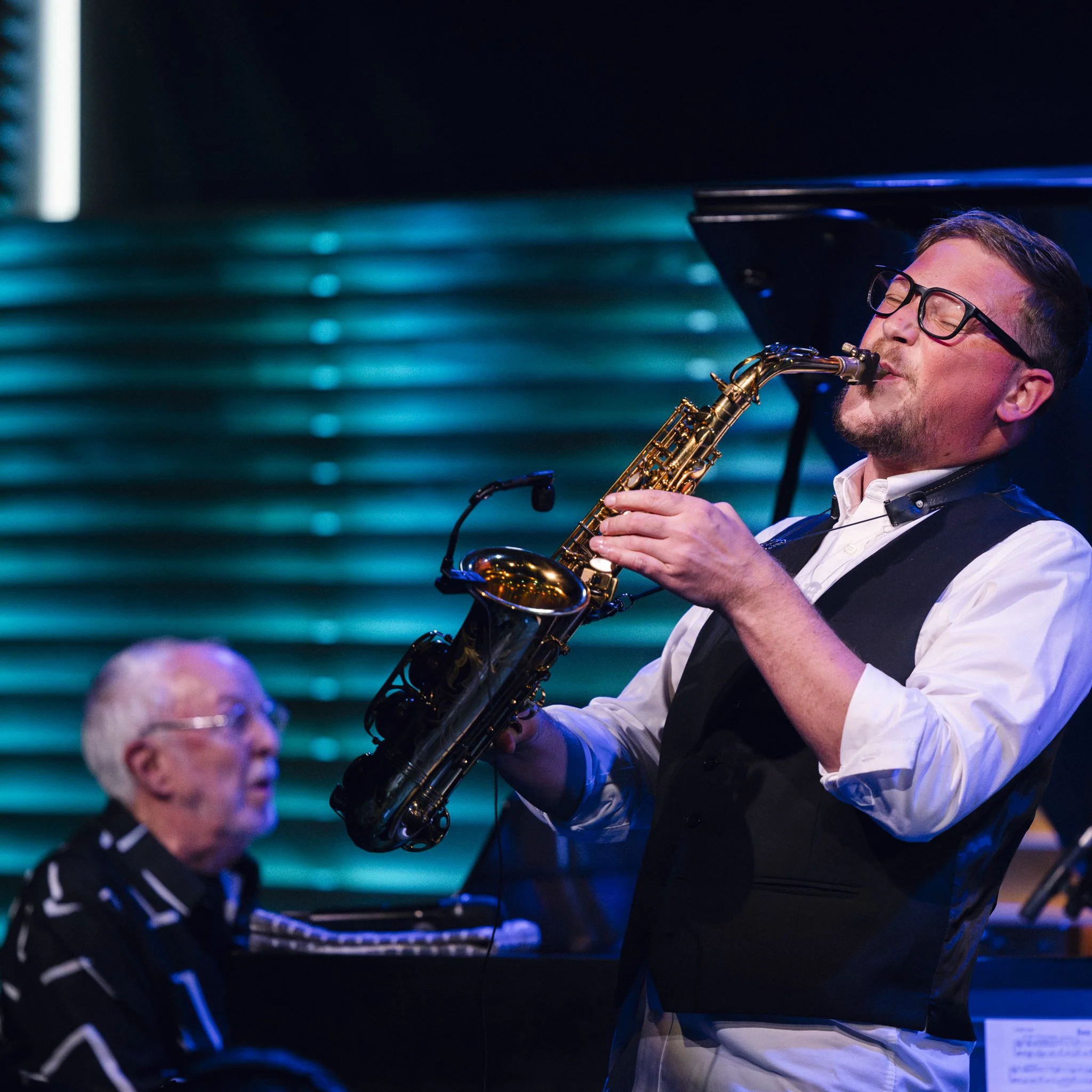 A man playing a saxophone on stage with a pianist in the background and a blue and black striped backdrop.