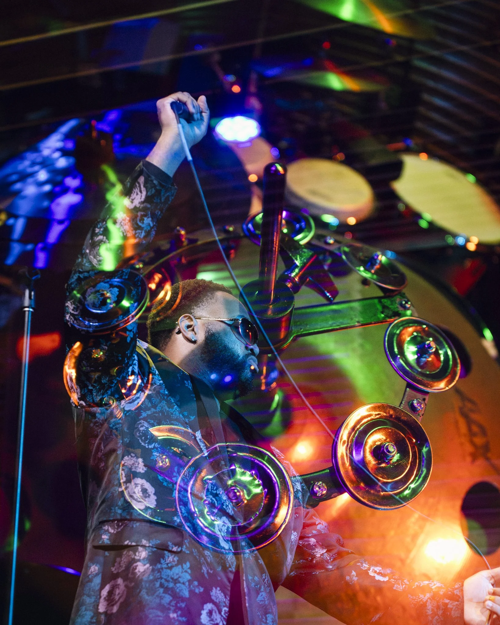 A DJ wearing sunglasses and a floral jacket performs with colorful lights and visuals in the background, holding a wire or cable.