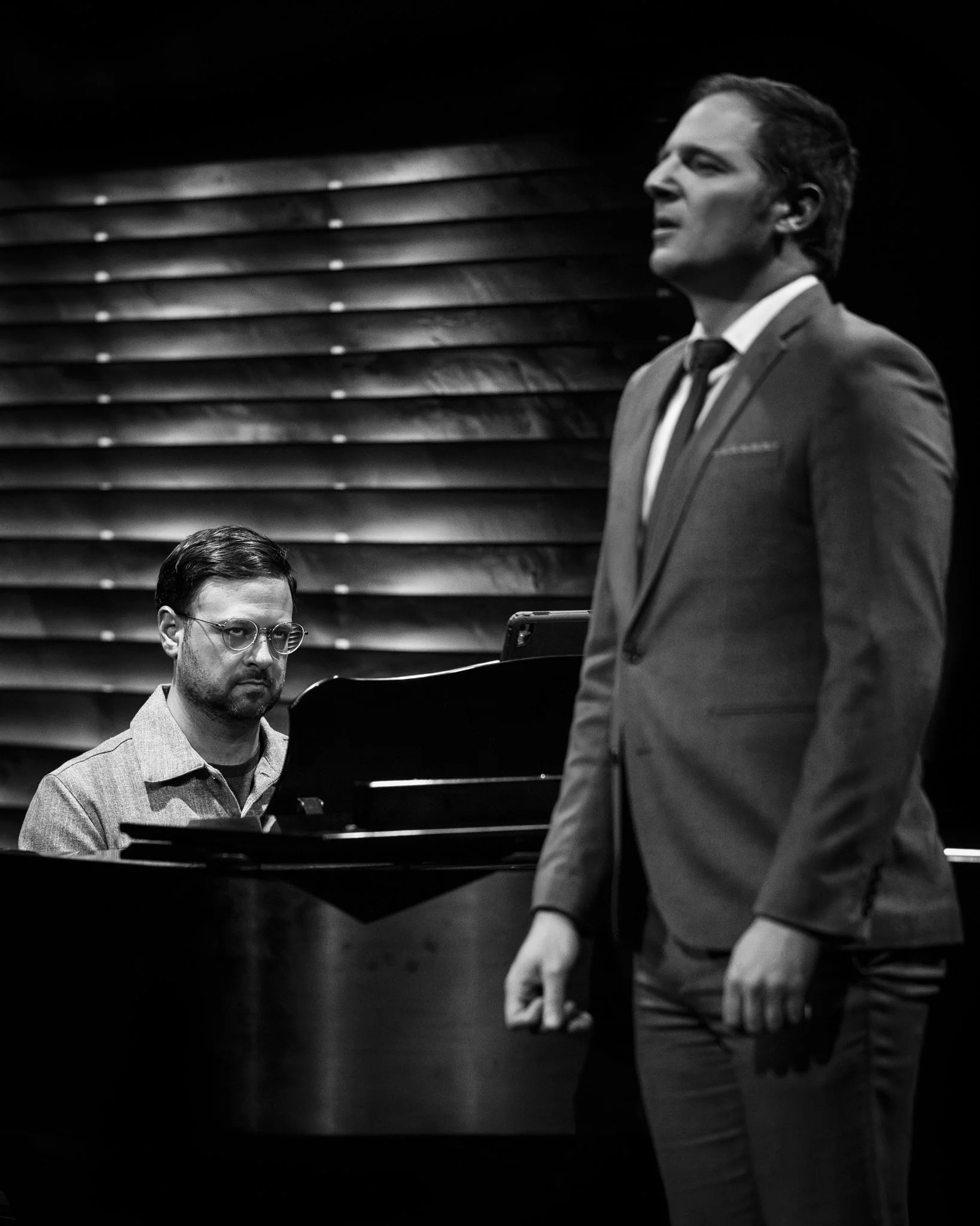 A black and white photo of a man in a suit singing, with a pianist in the background at a piano, on a dark stage.