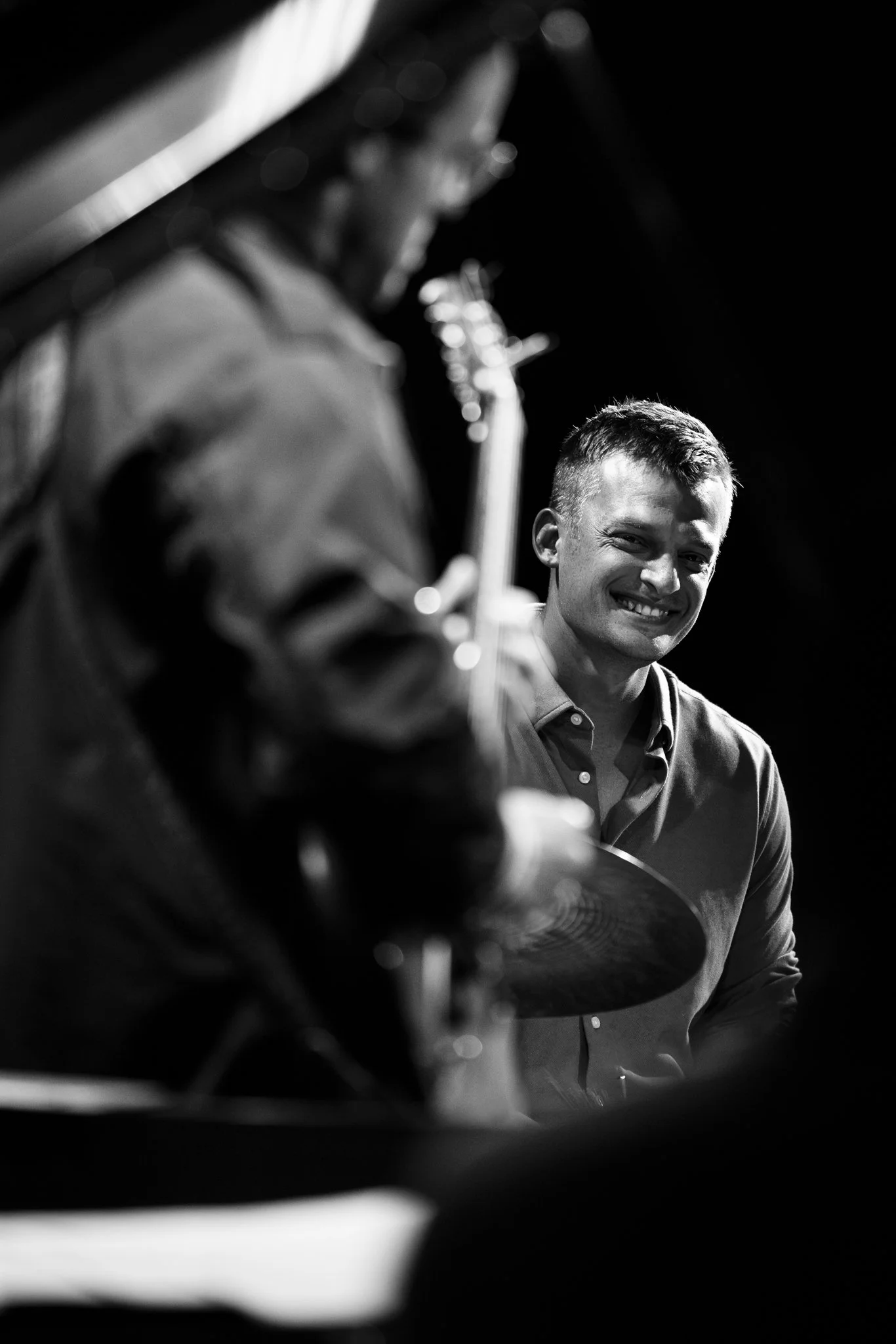 Black and white photo of a smiling man playing the drums, with a blurred person playing a guitar in the foreground.