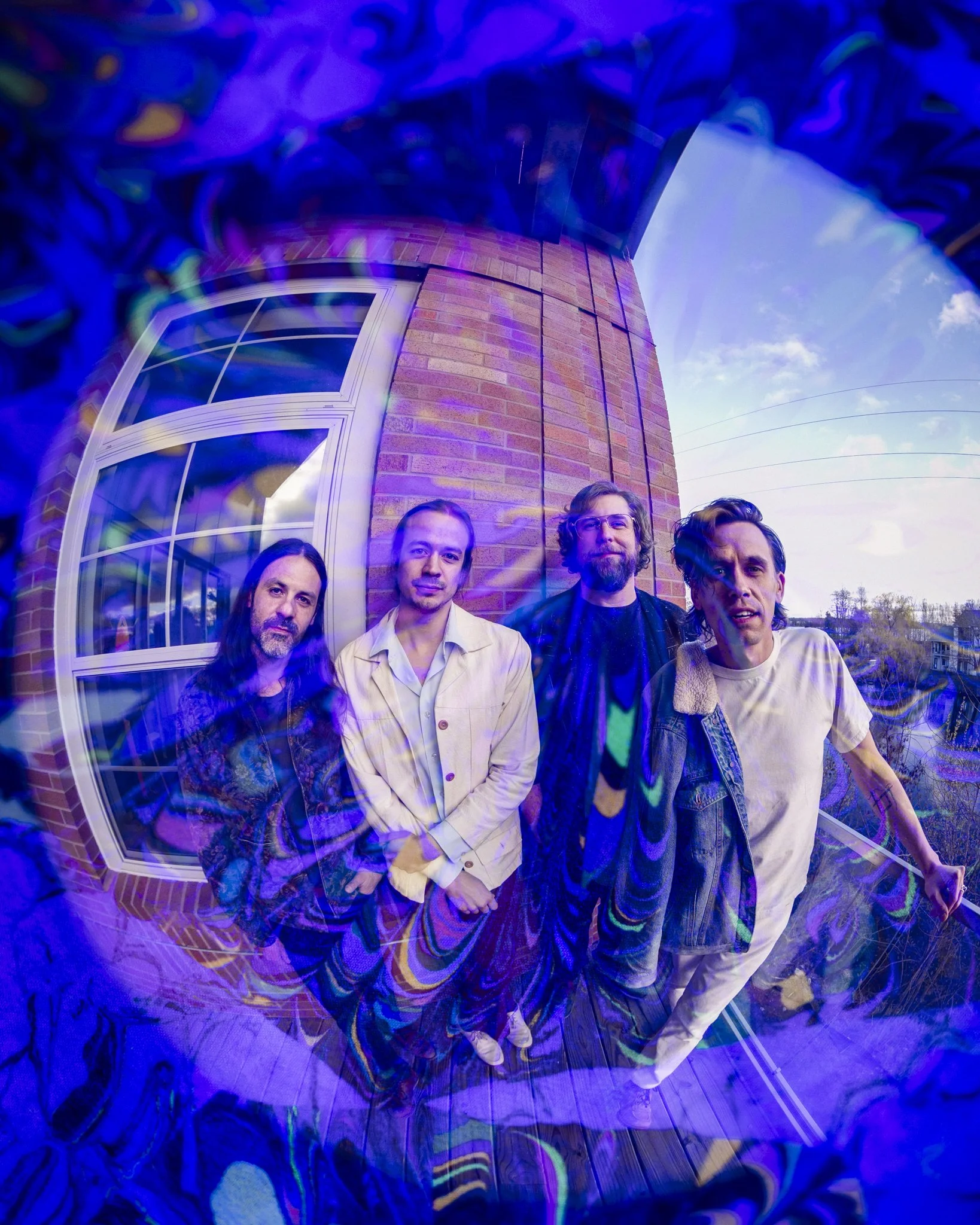 Four men standing on a wooden deck in front of a brick building with a window, captured with a fish-eye lens creating a distorted, colorful effect.