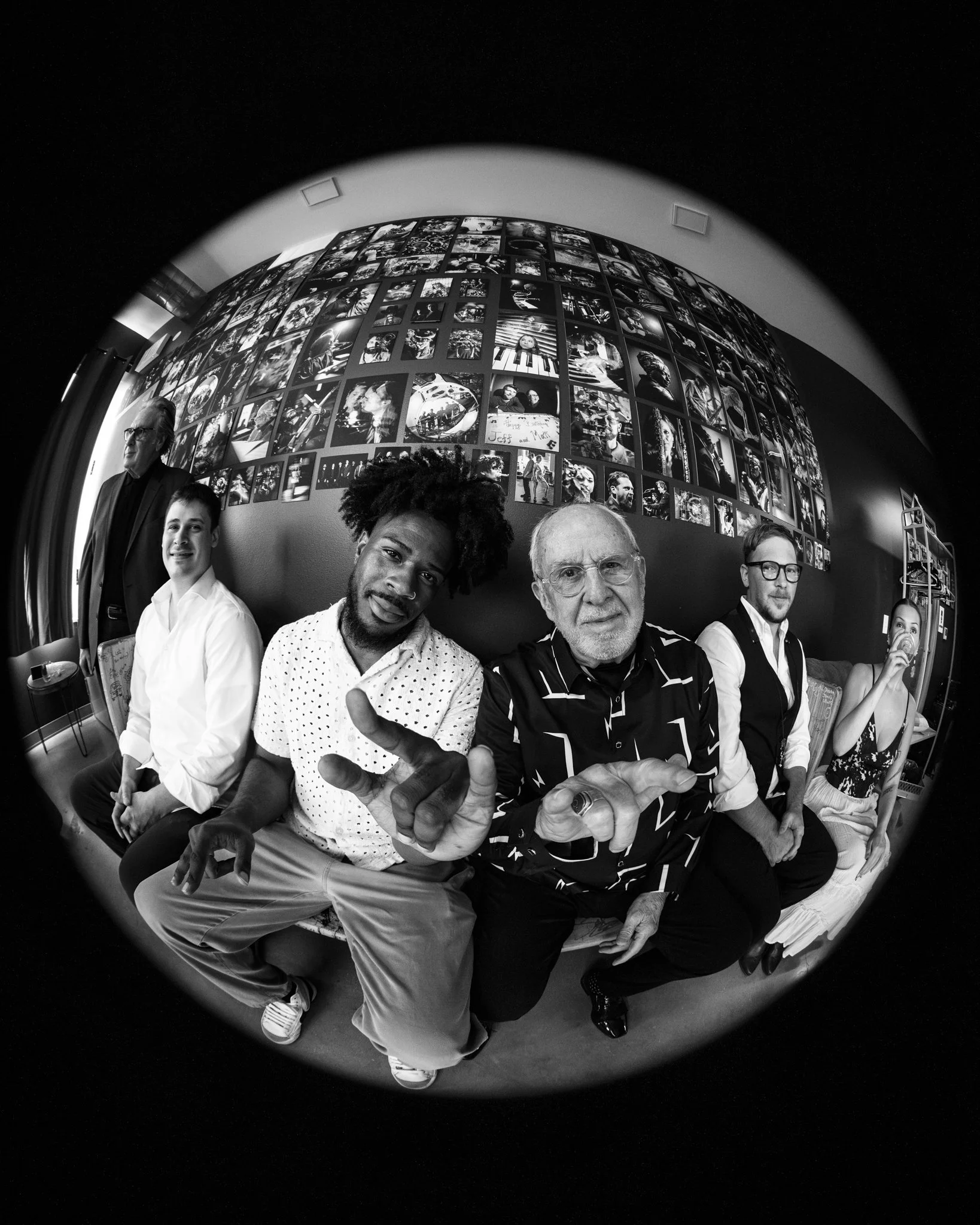 Black and white fisheye photo of six people sitting on a couch in a room with a wall covered in numerous photographs.