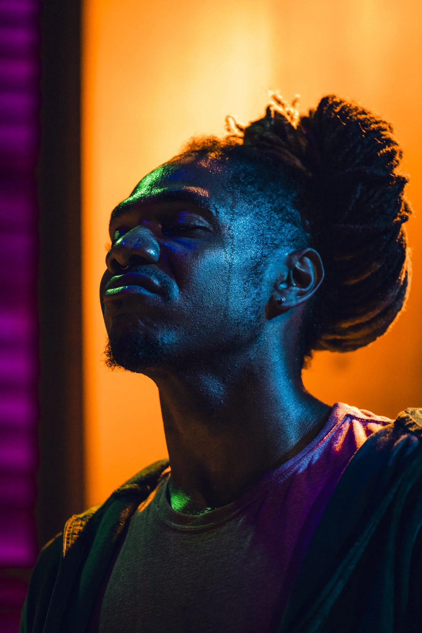 A young person with dreadlocks and a goatee, standing with eyes closed against a background with colorful lighting, illuminated by vibrant hues of orange, green, and purple.