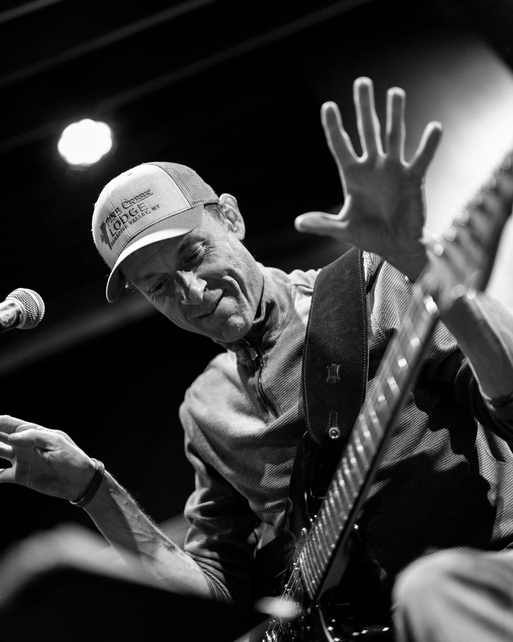 A man playing an electric guitar on stage, wearing a cap and smiling with his eyes closed, with a microphone nearby.