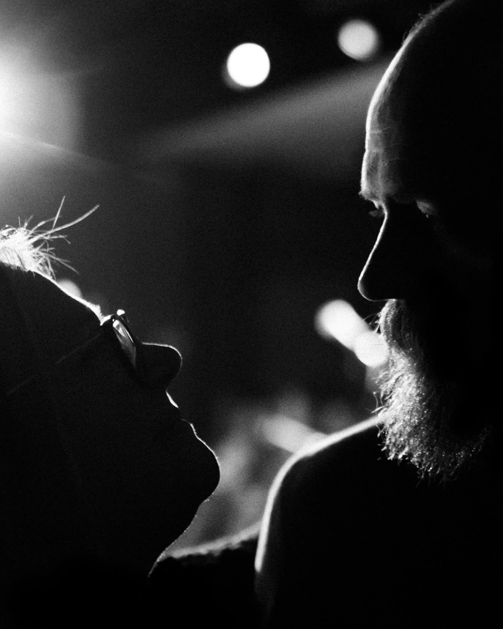 A black and white silhouette of two people facing each other closely, with one person wearing glasses and the other having a beard and bald head, in a dimly lit environment.