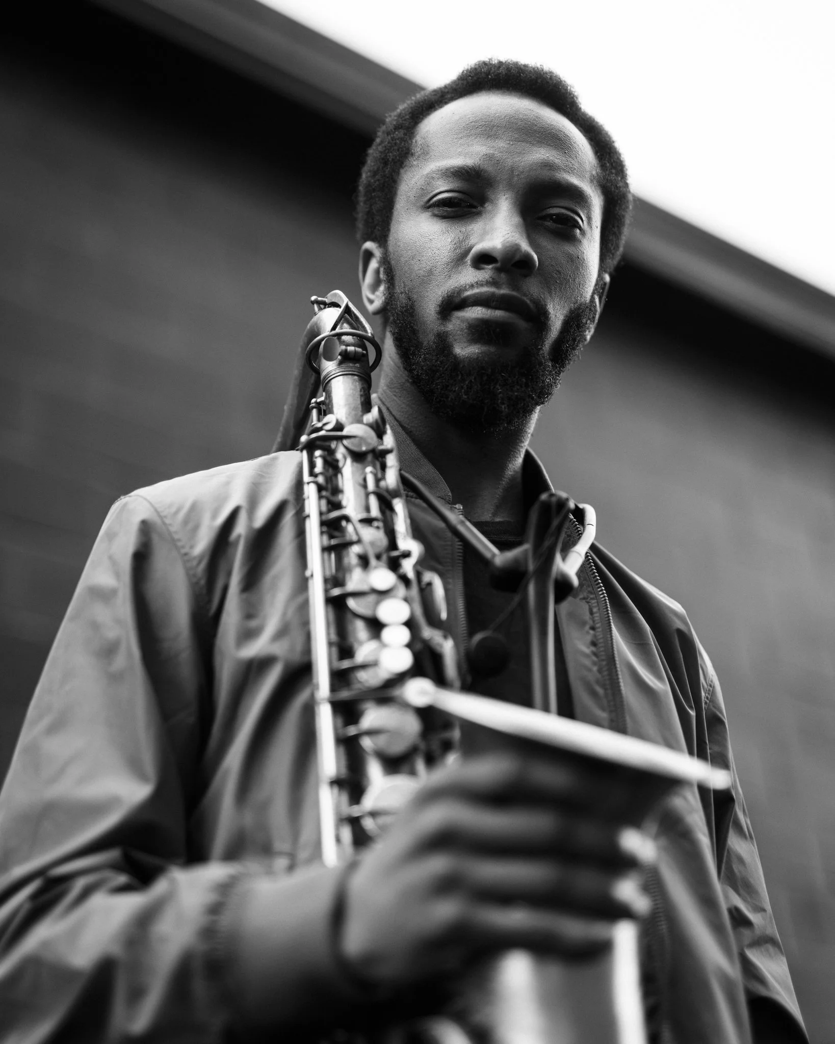 Black and white photo of a man holding a saxophone, looking directly at the camera, standing outdoors against a blurred background.