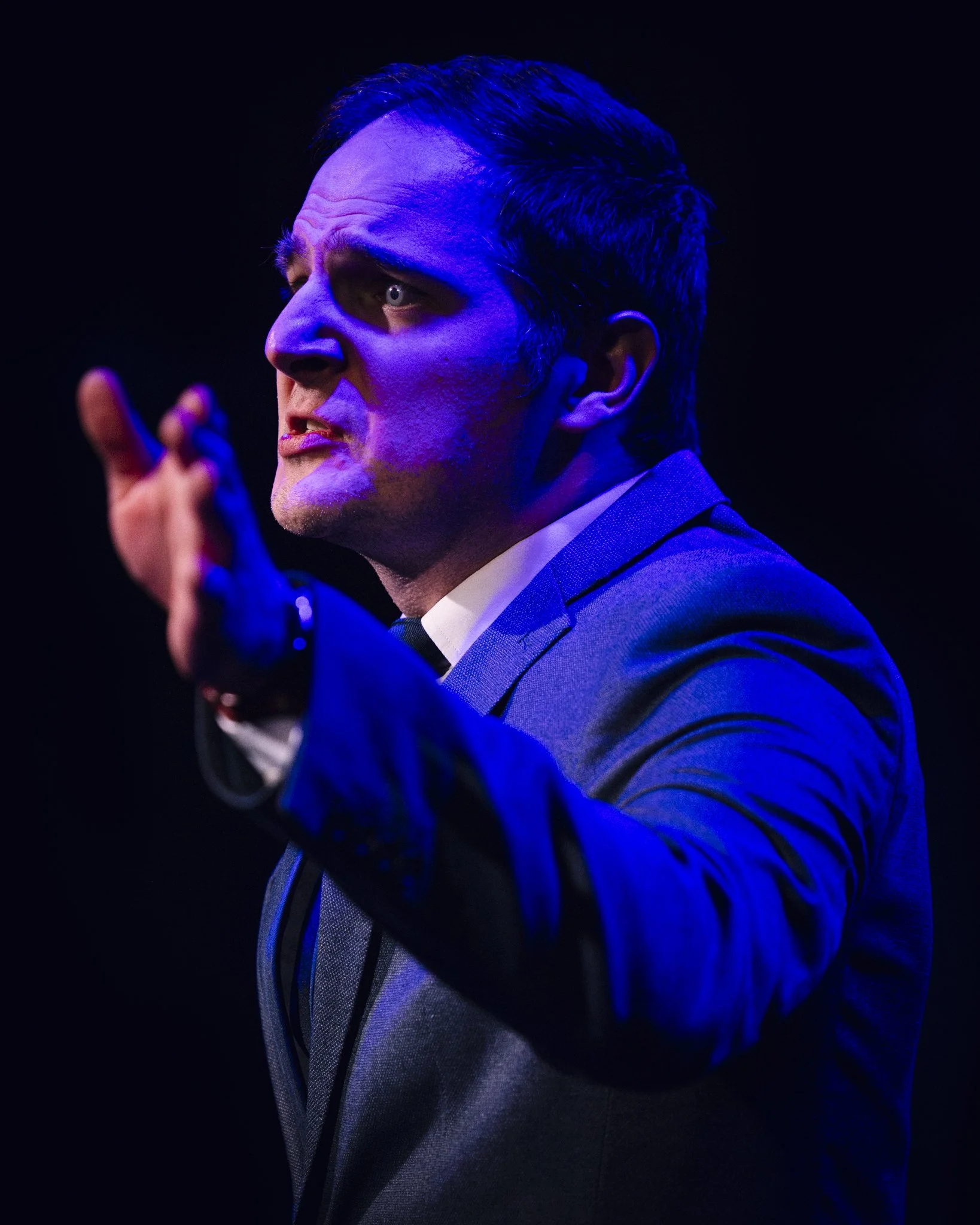 Man in suit making expressive gesture during a performance with dramatic lighting.