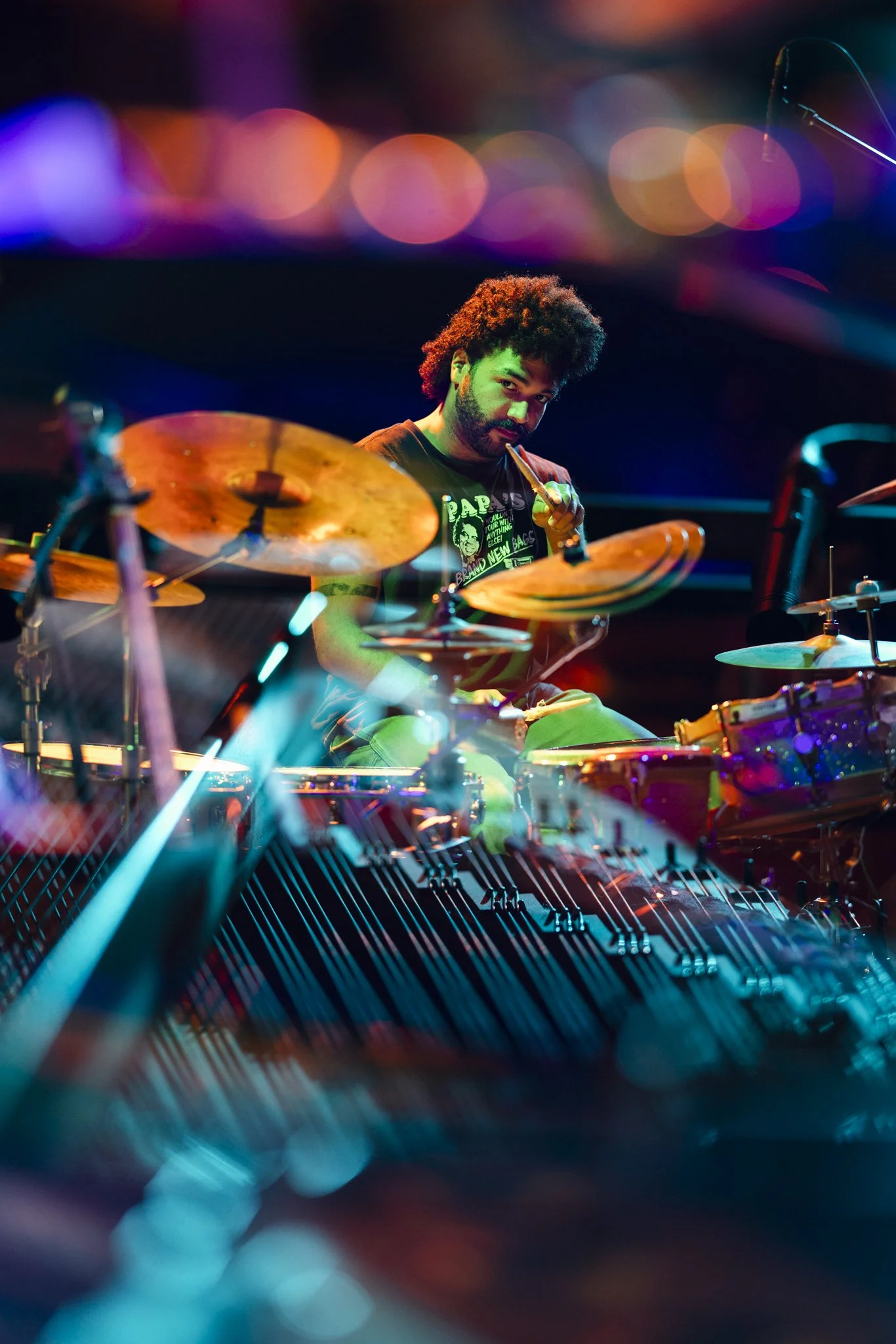 A man with curly hair playing a drum set on stage, illuminated by colorful lights, with a focus on the cymbals and drums.