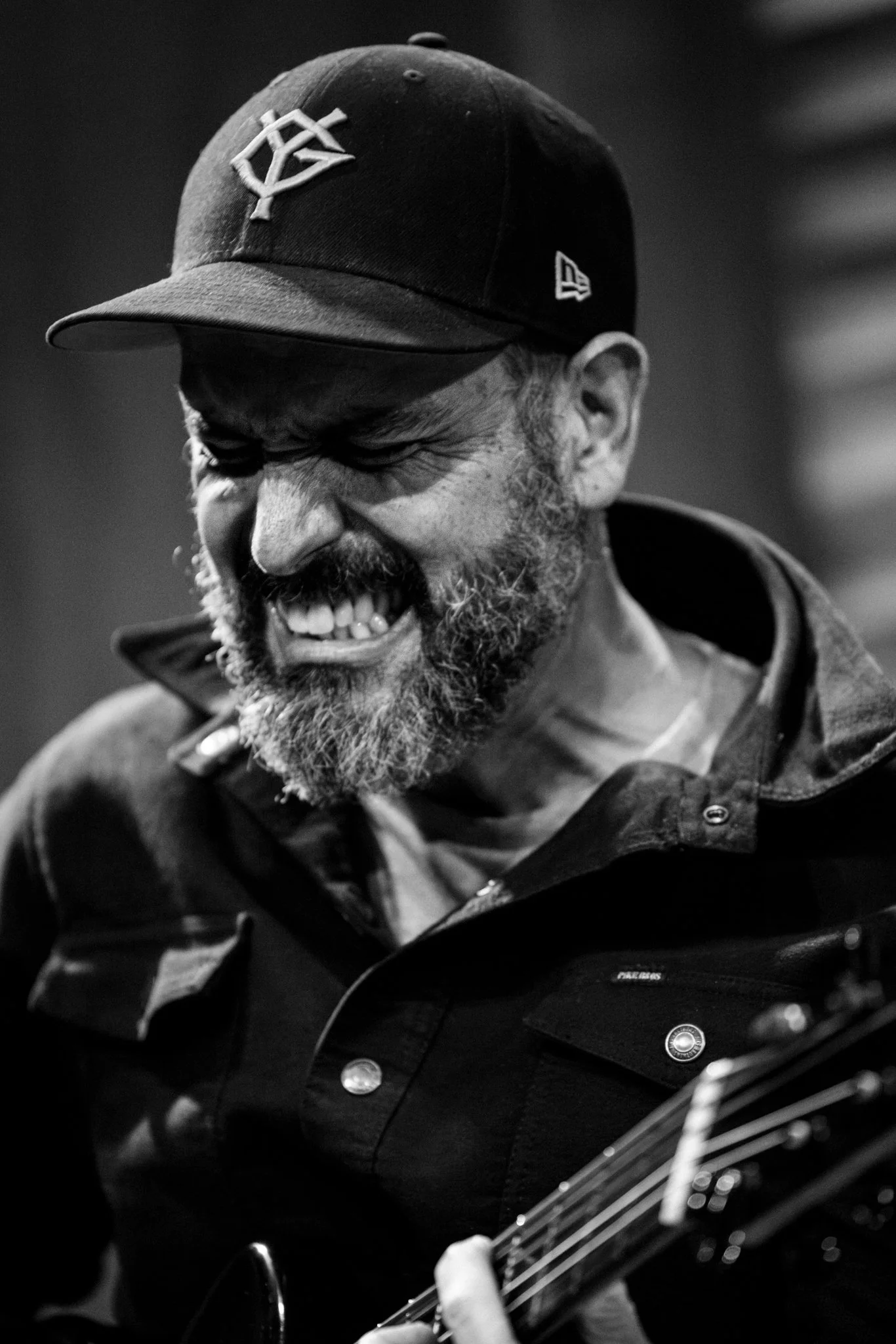 A black and white photo of a man with a beard and mustache, wearing a baseball cap and a jacket, smiling while playing an electric guitar.