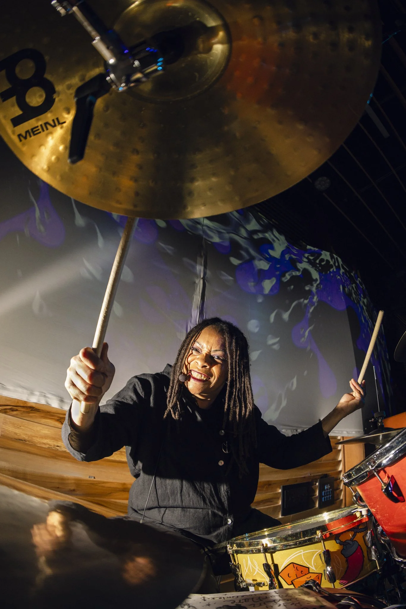 A woman with dreadlocks playing drums, smiling, and holding drumsticks in a lively setting with colorful background visuals.