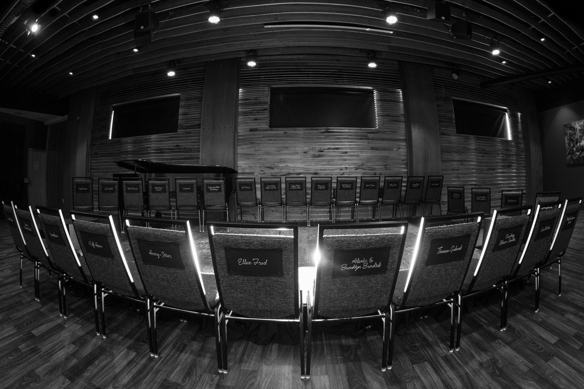 A black and white photo of an empty seating area with chairs labeled with names, arranged in a semi-circle facing a stage with a piano. The room has wooden walls and a wooden ceiling, with three windows on the wall behind the stage and dim lighting.