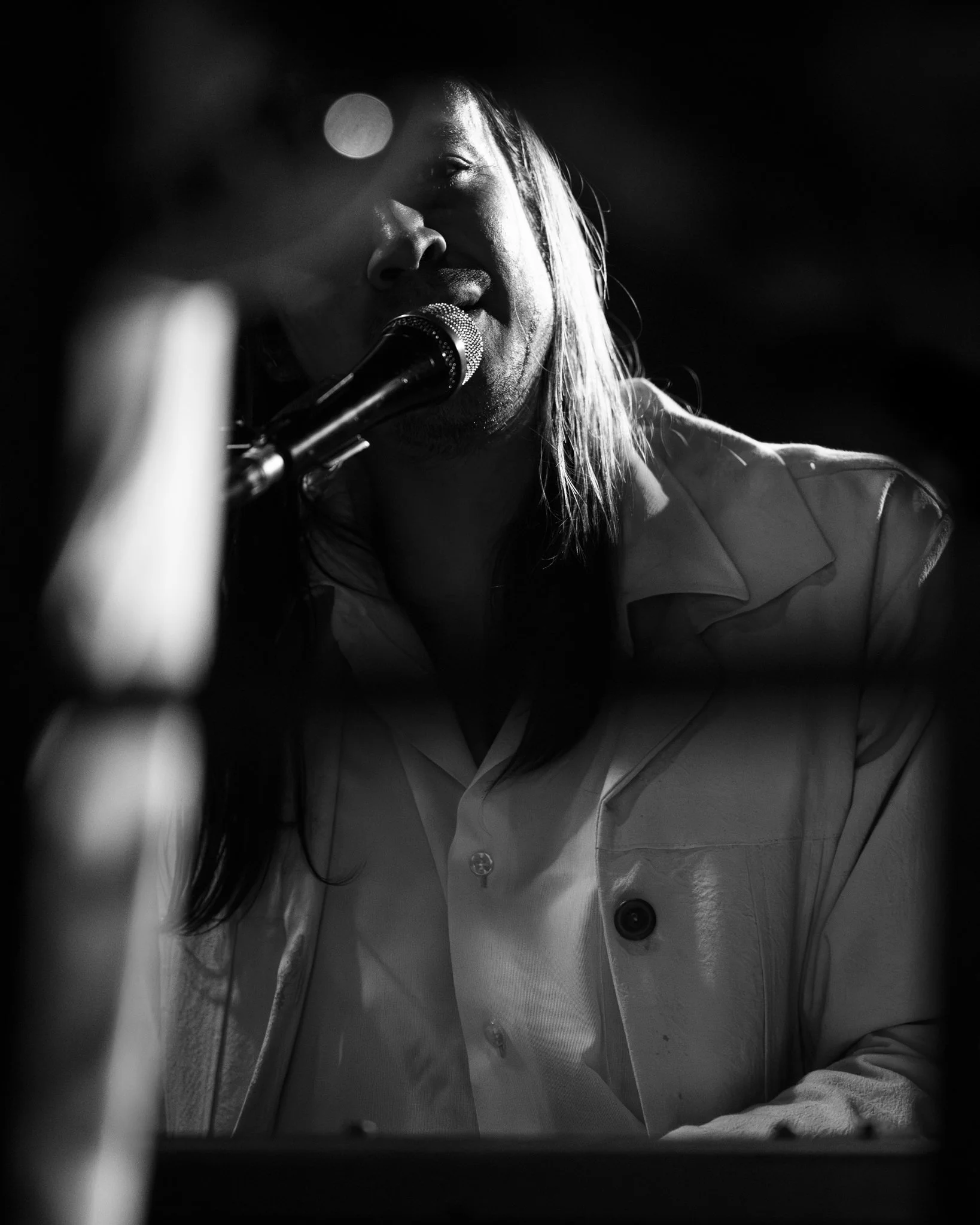 Black and white photo of a woman singing or speaking into a microphone, with light reflecting on her face and hair, wearing a button-up shirt.