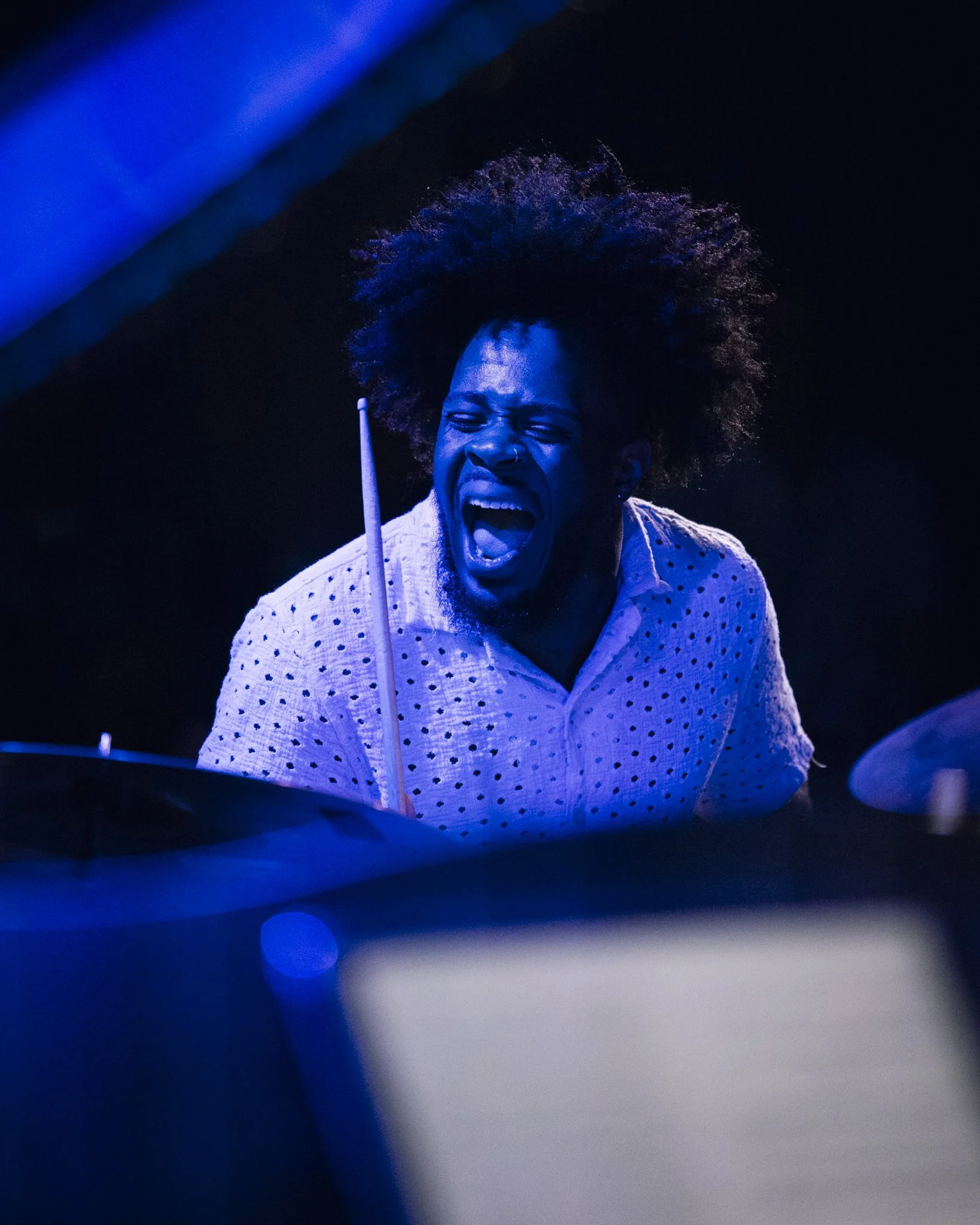 A man with curly hair passionately playing drums, wearing a white shirt with black spots, illuminated by blue light.