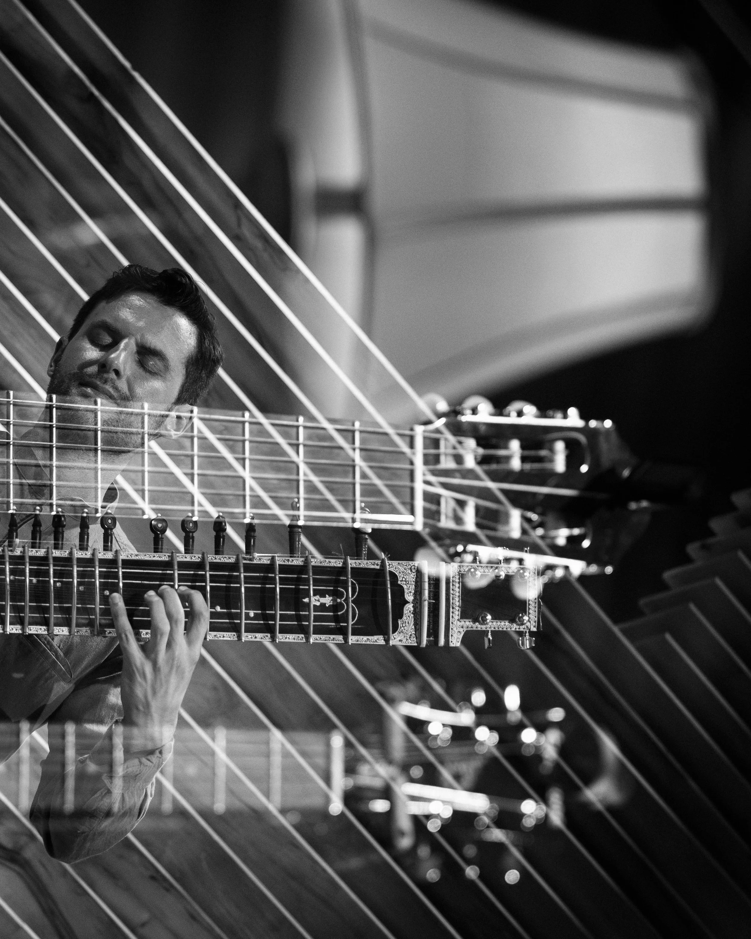 A black and white photo of a man playing a guitar, with his eyes closed, possibly deeply immersed in music. The man is shown leaning his head against the fretboard of the guitar.