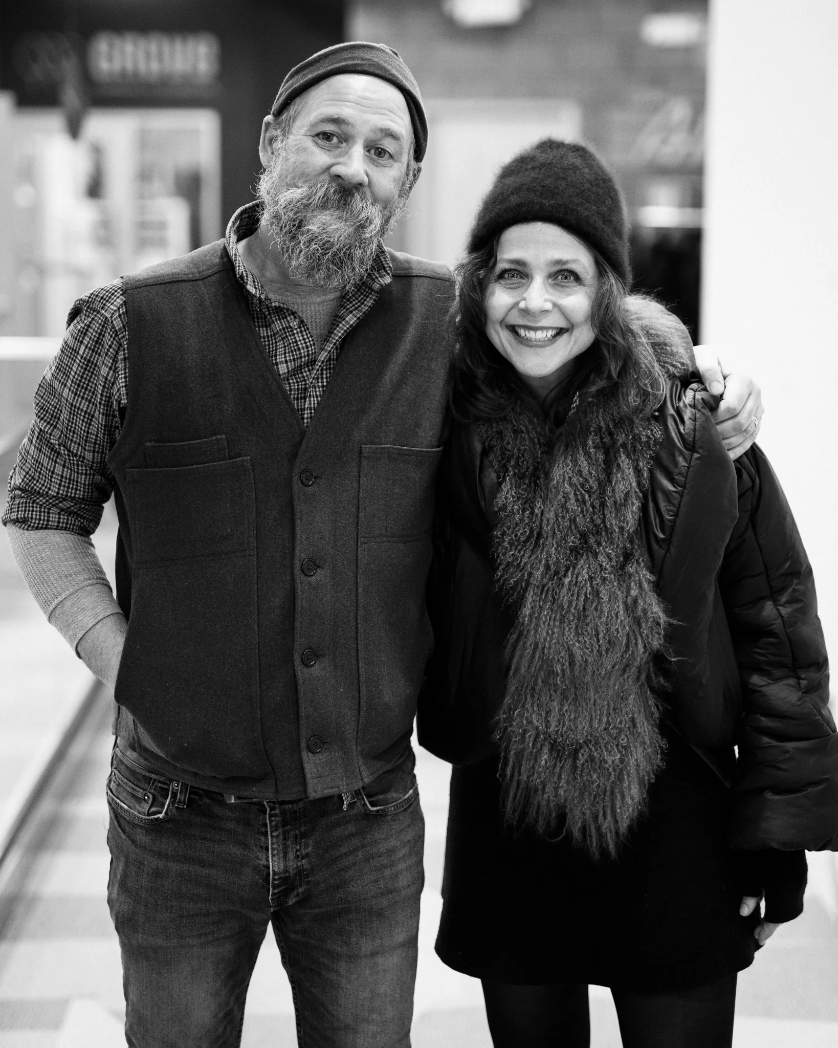 A black and white photo of a man and a woman smiling, standing close together, with the man’s arm around the woman’s shoulder. The man has a beard and is wearing a beanie, a checkered shirt, and a vest. The woman has curly hair and is also wearing a 