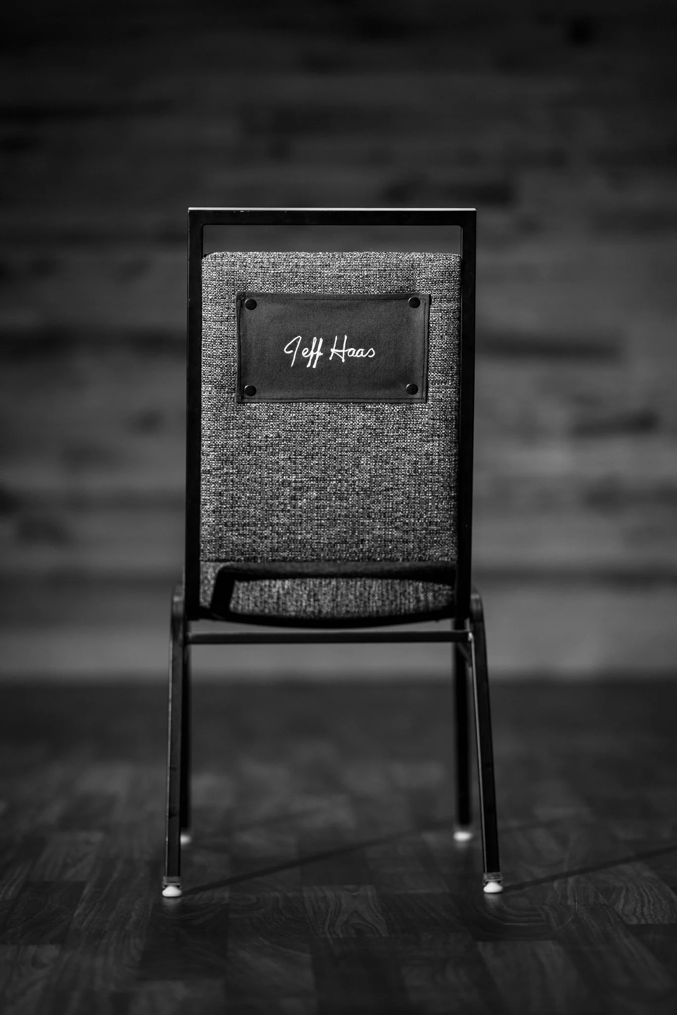 A black and white photo of a chair with a nameplate that reads 'Jeff Haas' hanging on its backrest.