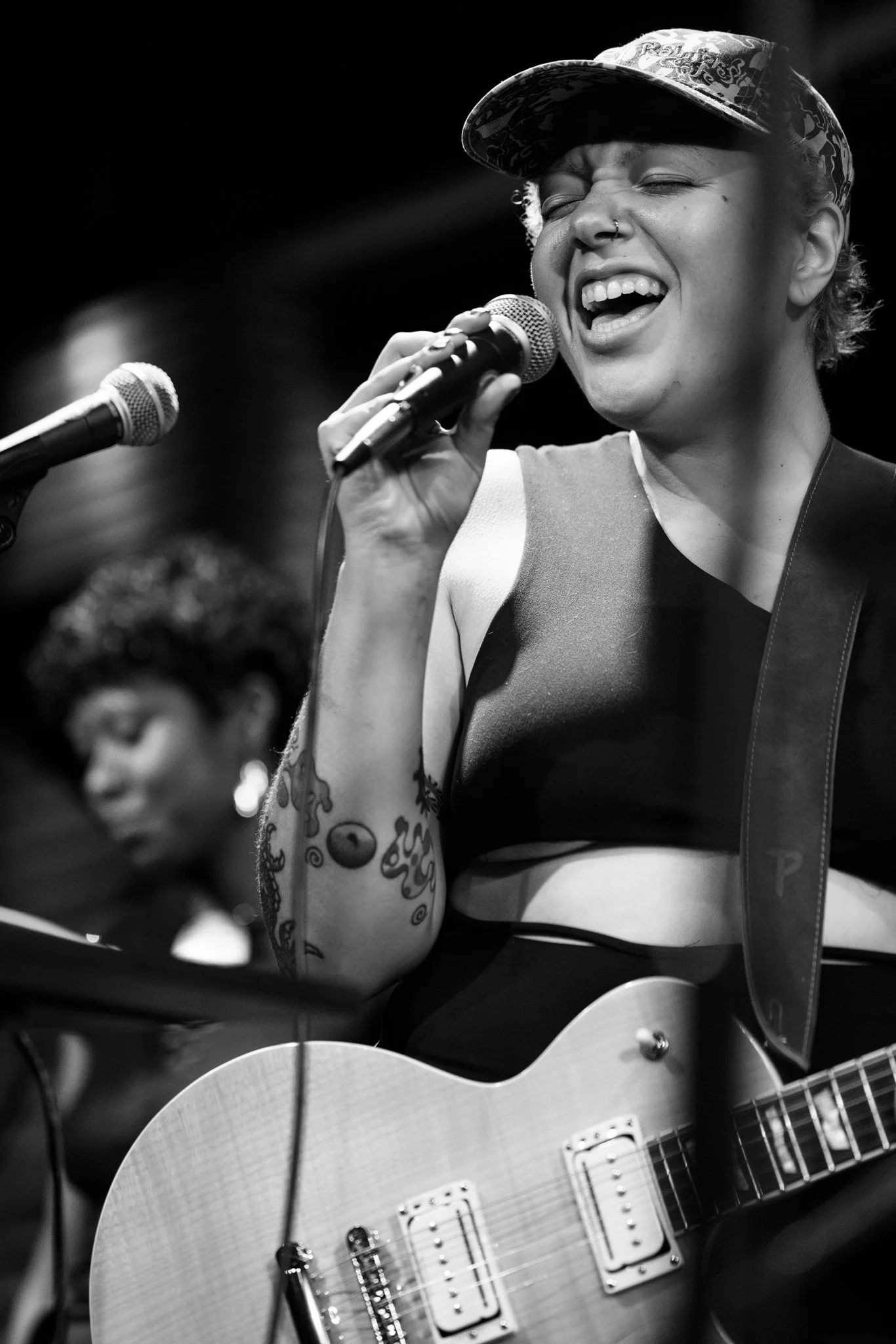 A woman singing into a microphone while playing an acoustic guitar, smiling with eyes closed, tattoos on her right arm, wearing a tank top and a cap, with a blurred person in the background.