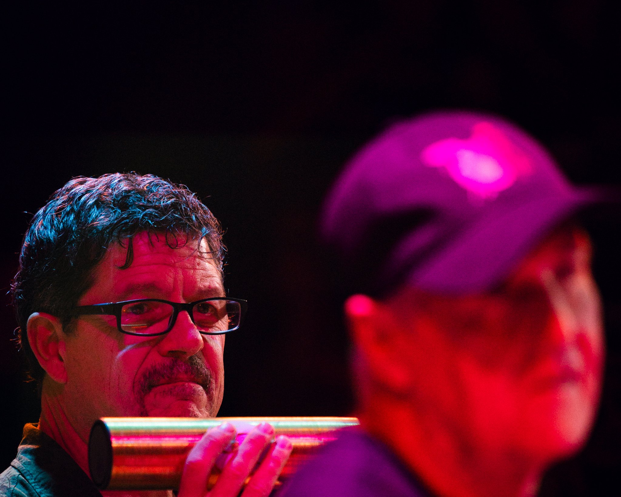 Two men, one with glasses and curly hair holding a colorful tubular instrument, the other wearing a cap, both in low lighting with pink and red hues.