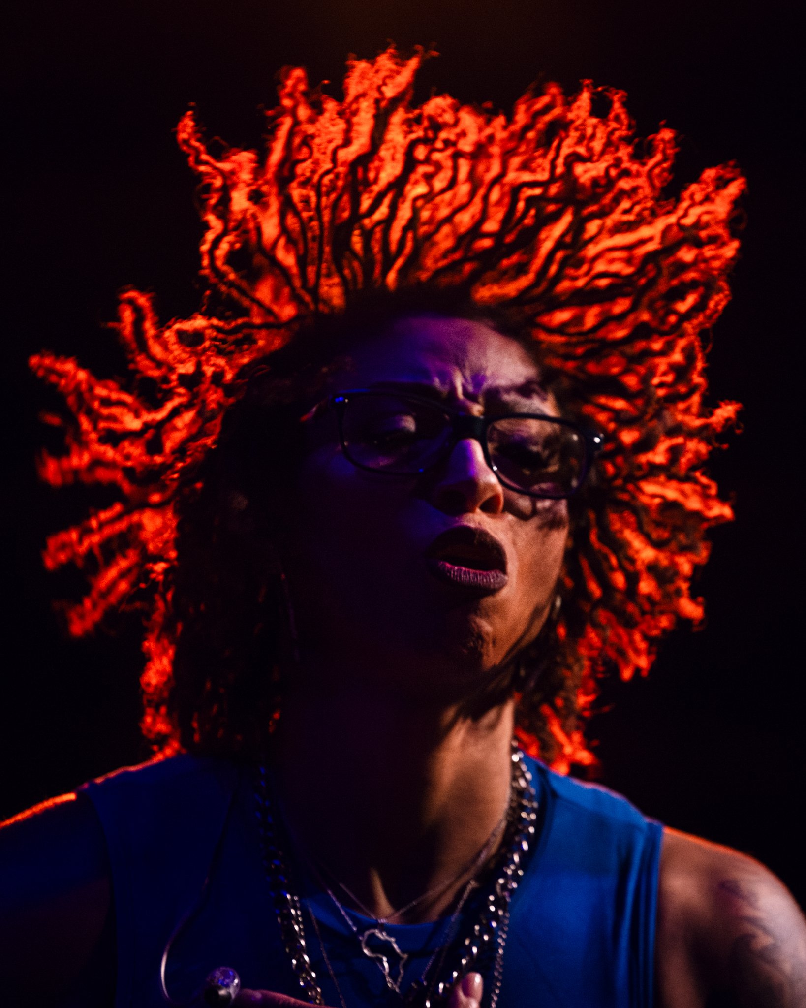 A person with long, fiery red dreadlocks, wearing glasses and dark makeup, lit dramatically against a dark background.