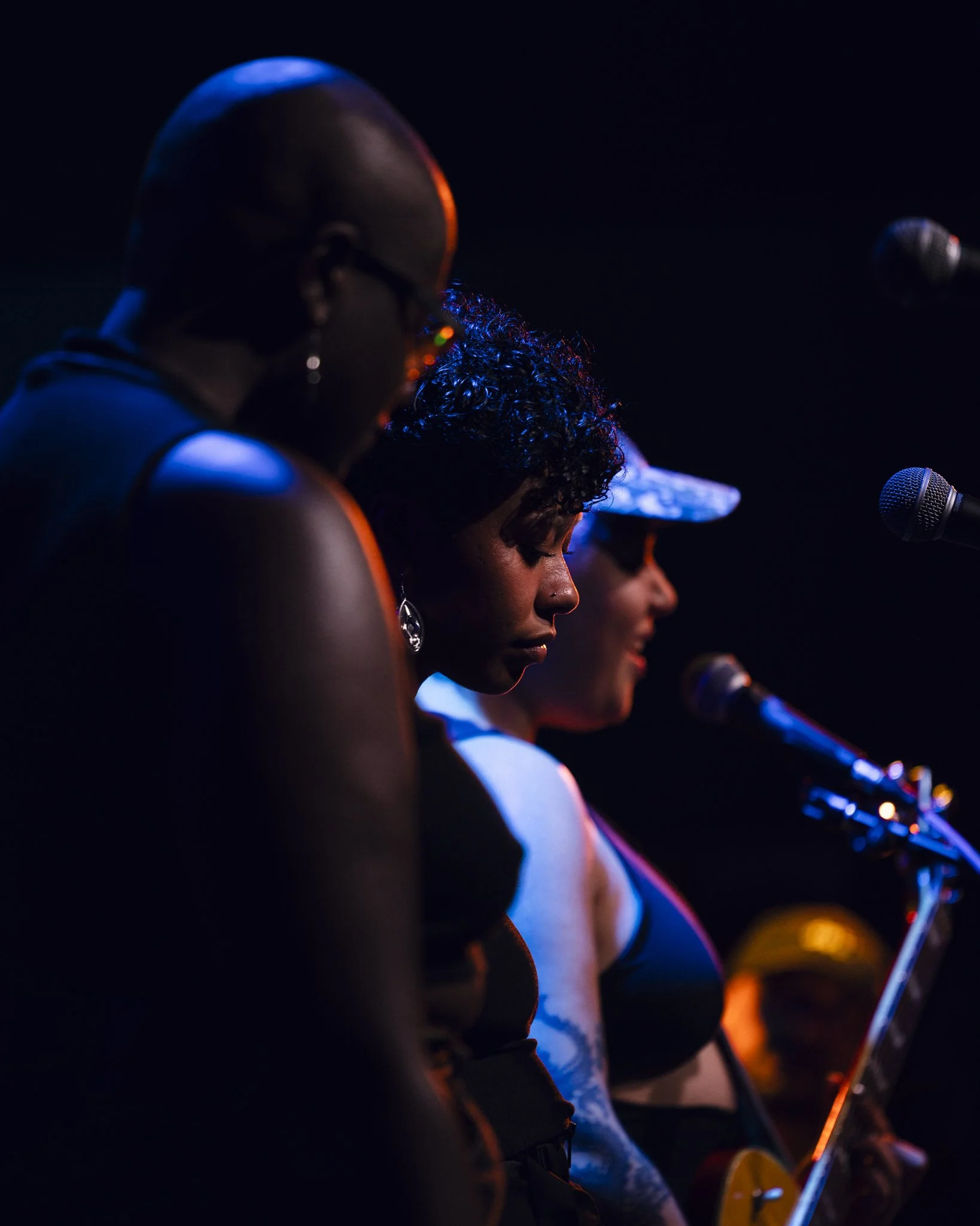 Three women singing or speaking into microphones on stage with blue and orange lighting.