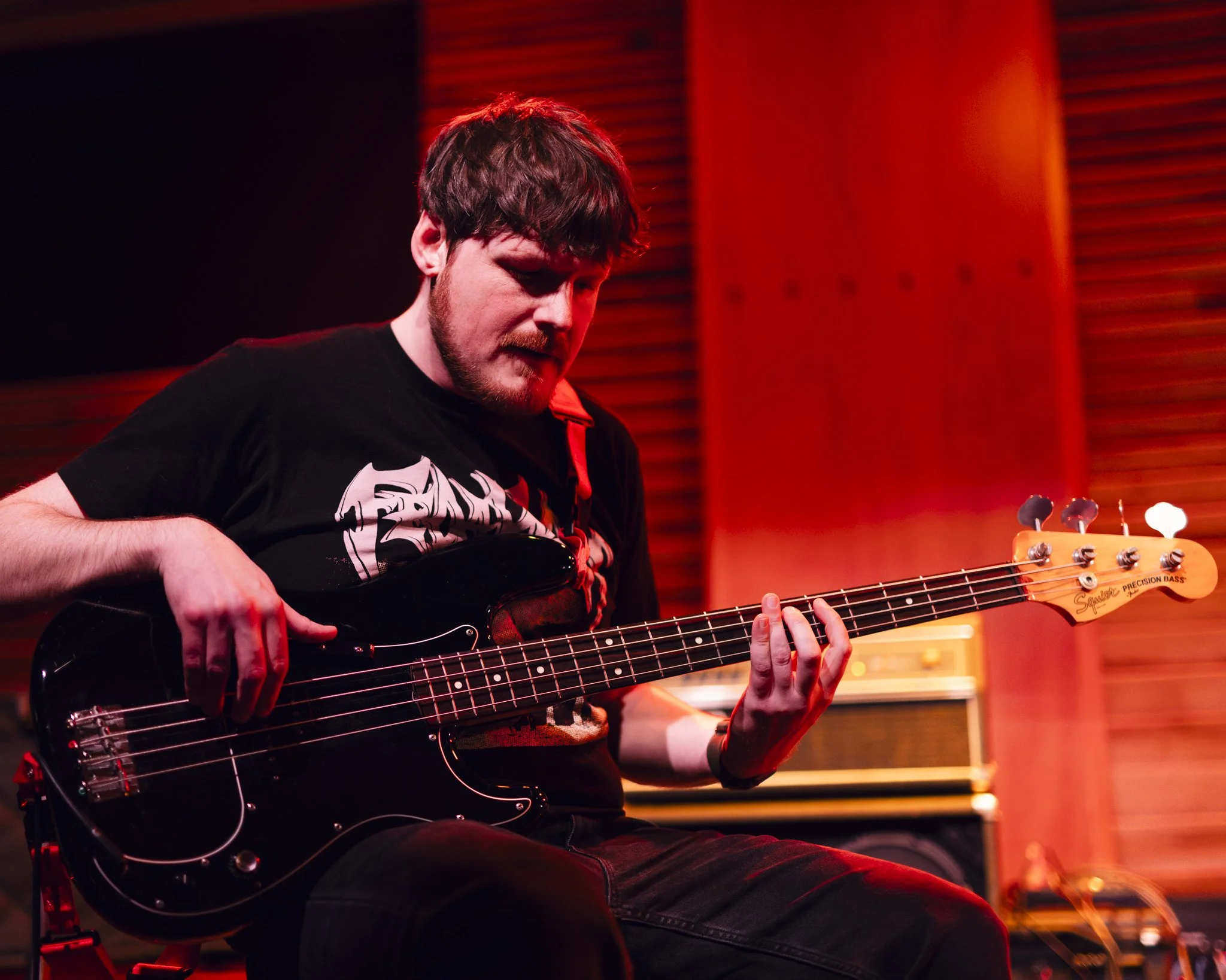A man playing a black electric bass guitar in a recording studio with warm lighting and wooden wall panels.