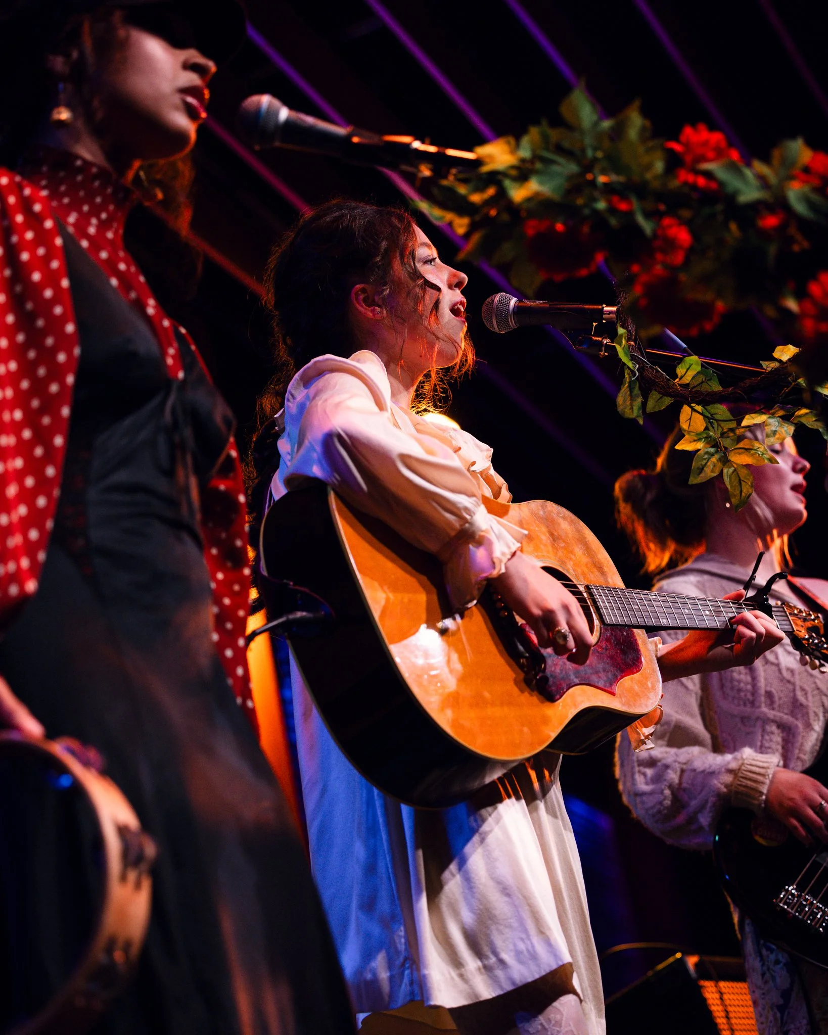 Three women performing on stage, one playing guitar, with colorful lighting and floral decoration overhead.