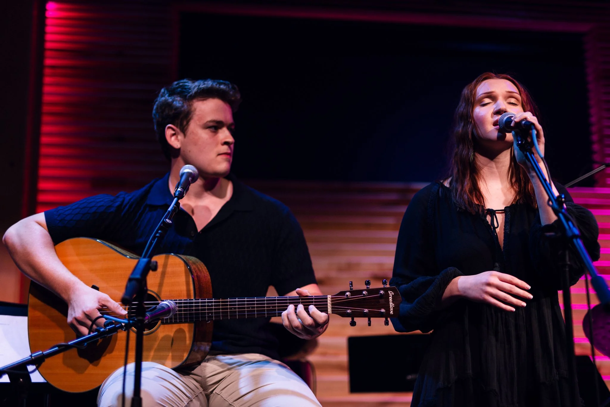 A young man playing an acoustic guitar and a young woman singing into a microphone during a live music performance.