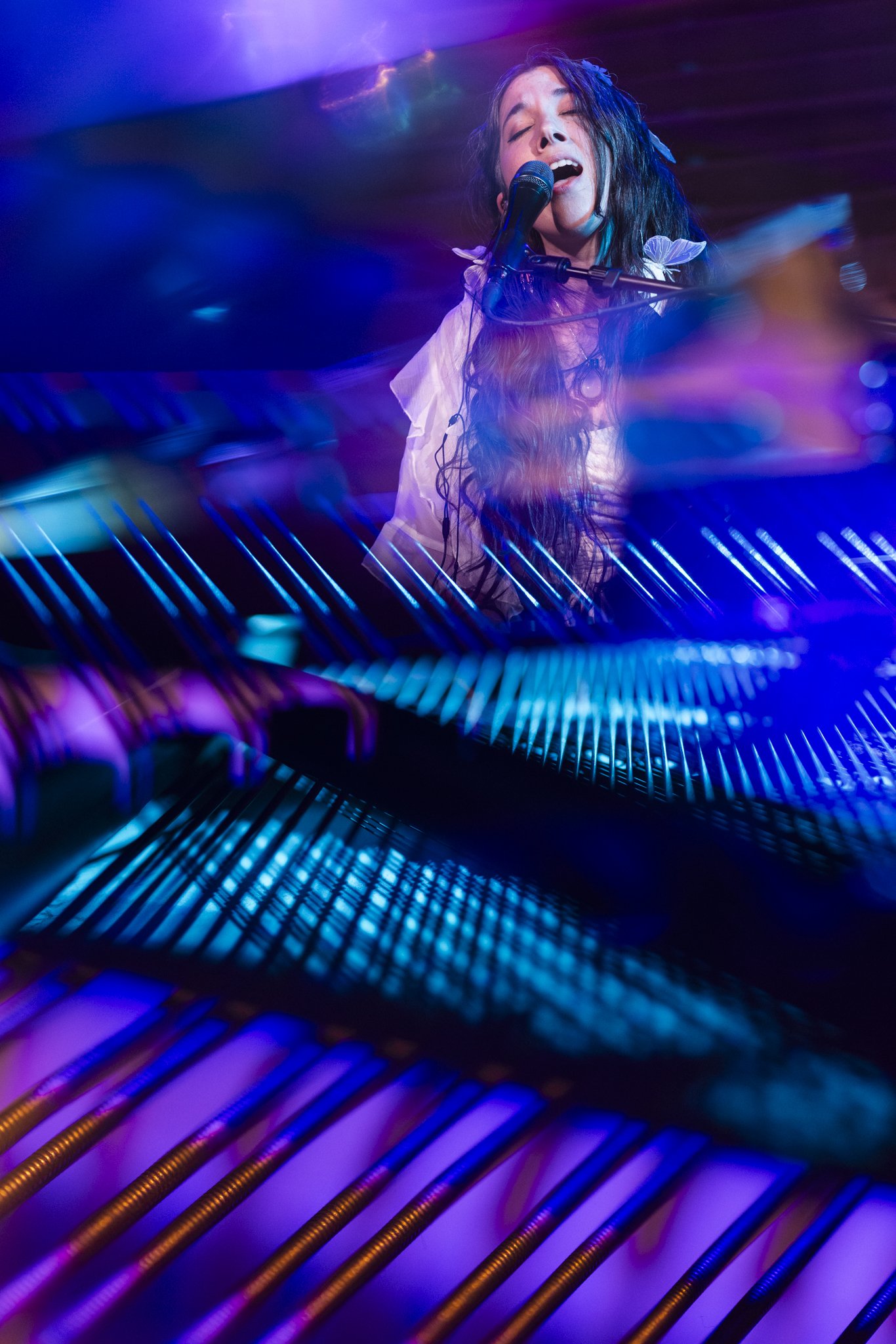 A woman singing passionately into a microphone while playing the piano, with colorful stage lights creating a vibrant atmosphere.