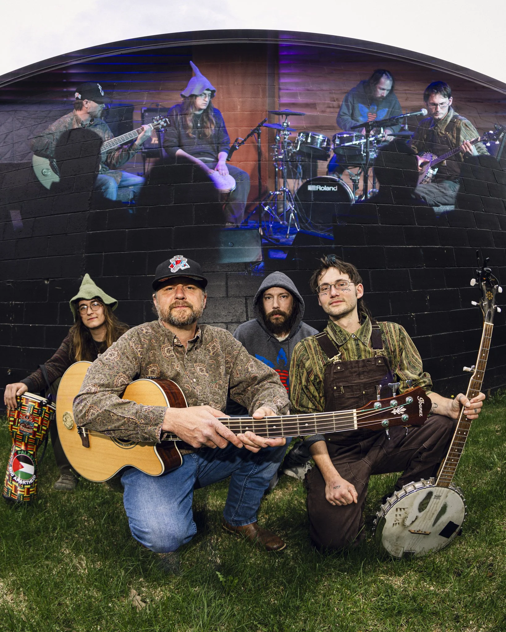 Group of five musicians outdoors, two with guitars, one with banjo, and one on drums, with an indoor performance scene projected illusion on the backdrop.